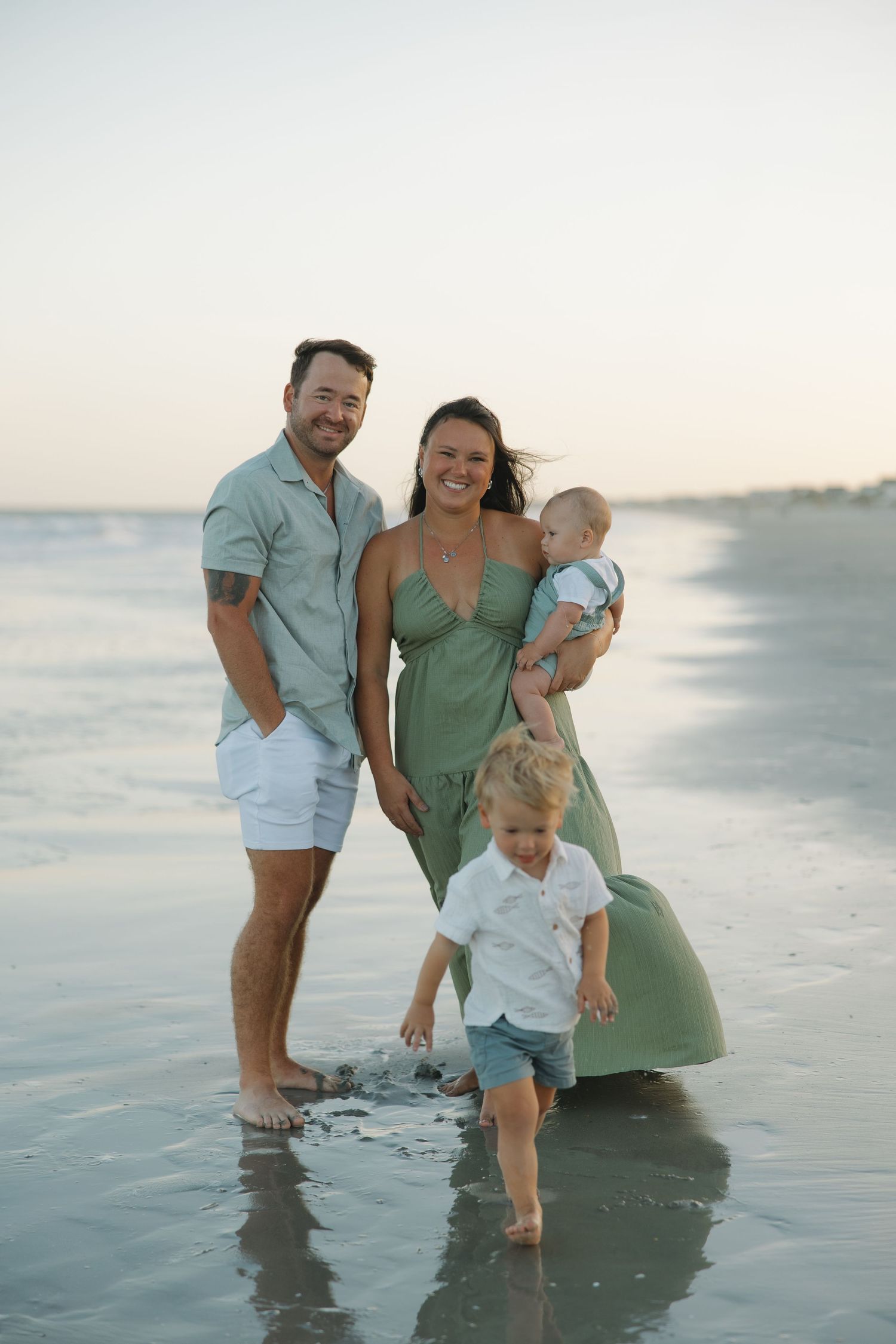 Family of four posing on beach at sunset, parents in green outfits with baby and toddler playing in shallow waves.
