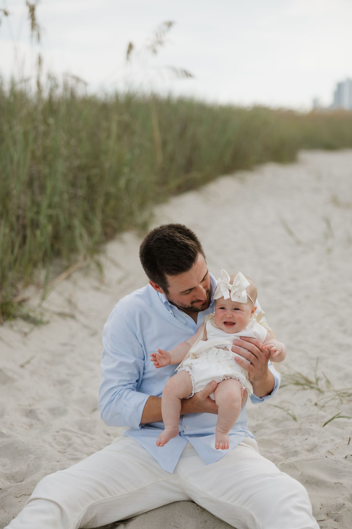 A father sits on sandy beach holding his smiling baby girl wearing a gold crown, with sea oats in the background.