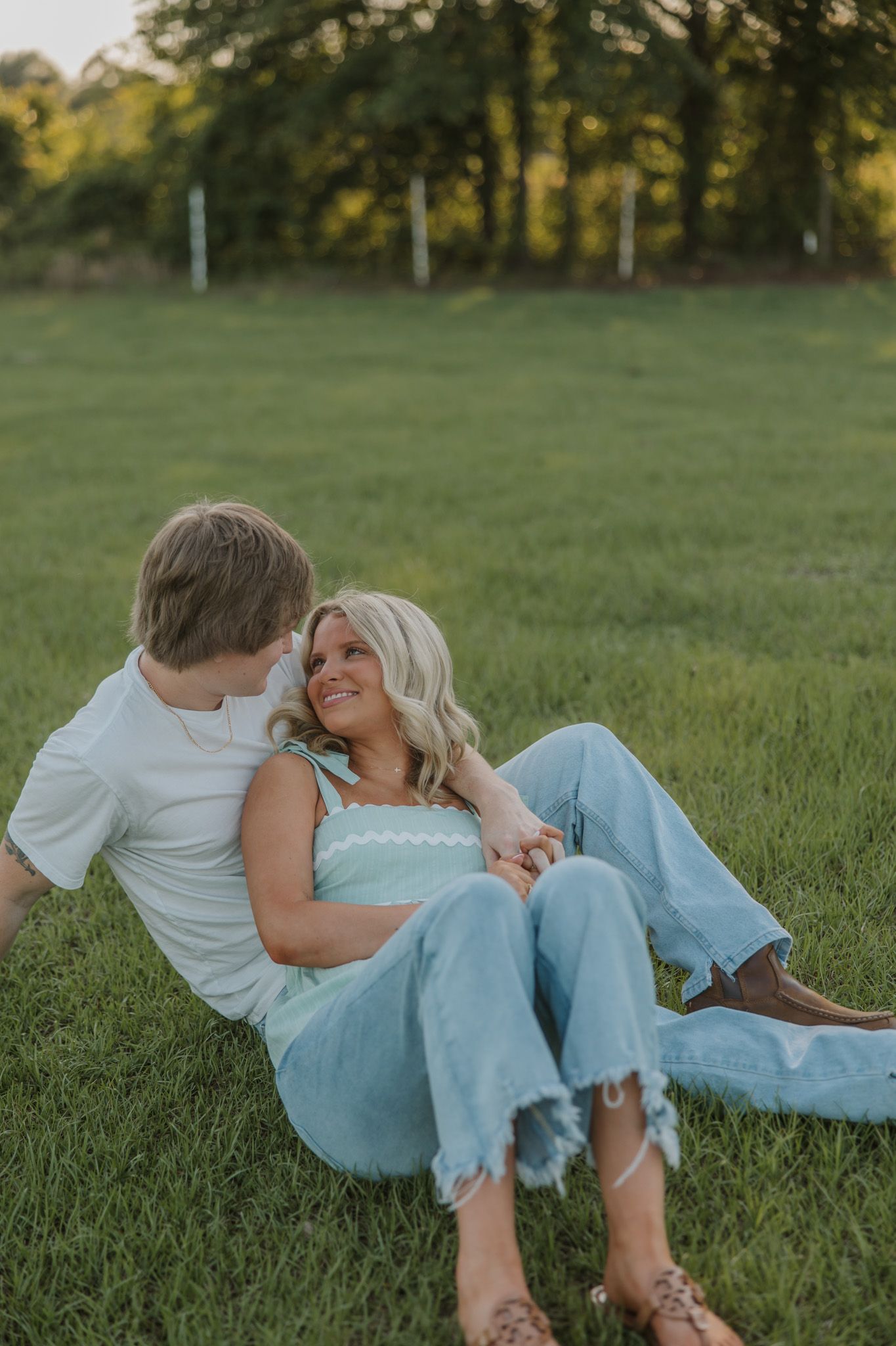 Couple sitting together on green grass field, woman in mint top and jeans smiling at man in white tee during golden hour.