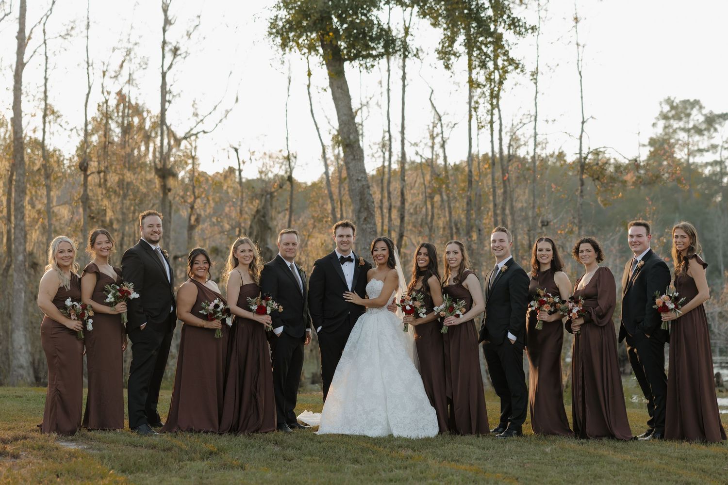 Bride and groom pose with their wedding party of bridesmaids in brown dresses and groomsmen in dark suits outdoors among tall trees.