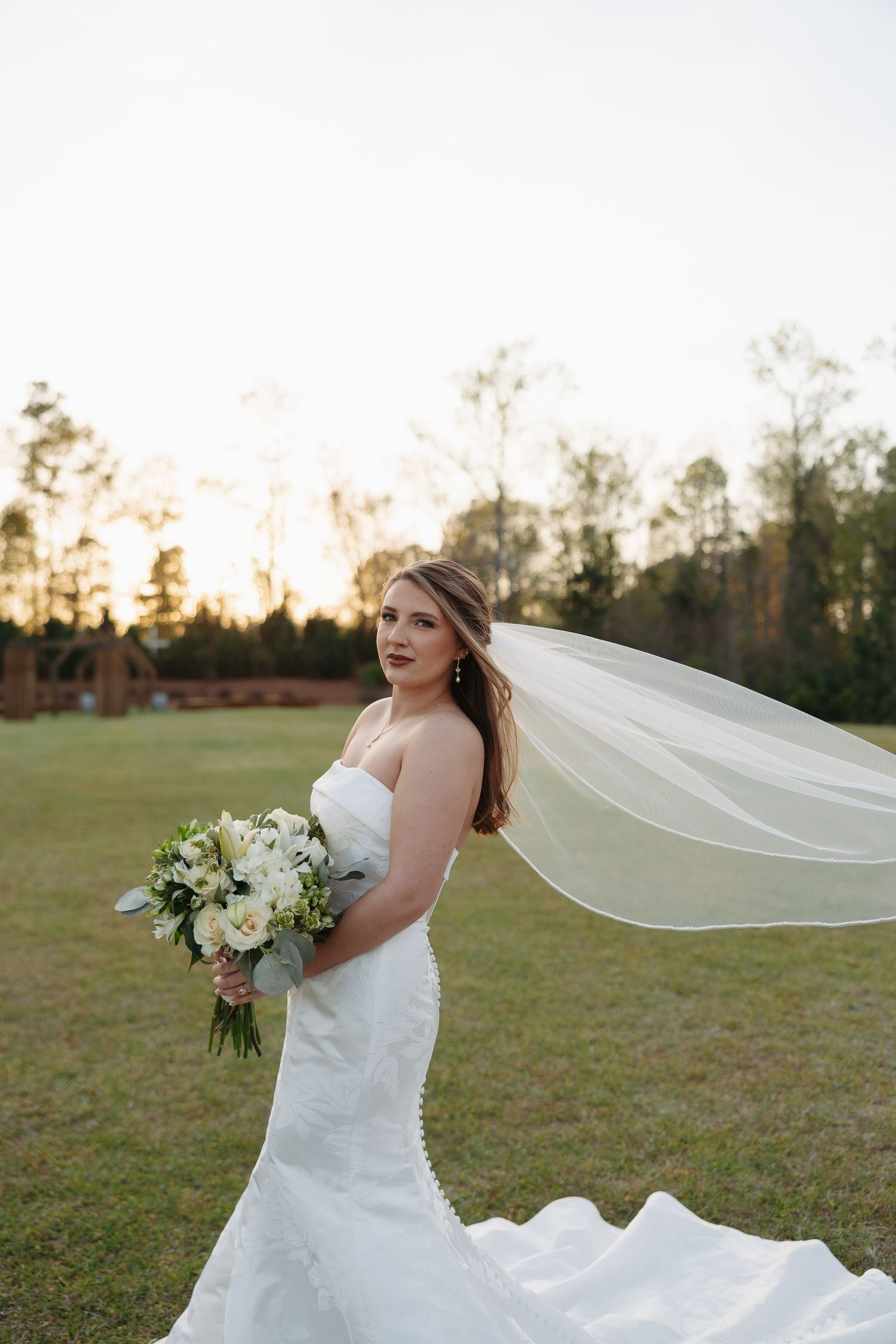 Bride in white strapless gown with flowing veil in wind holds white bouquet outdoors at golden hour.