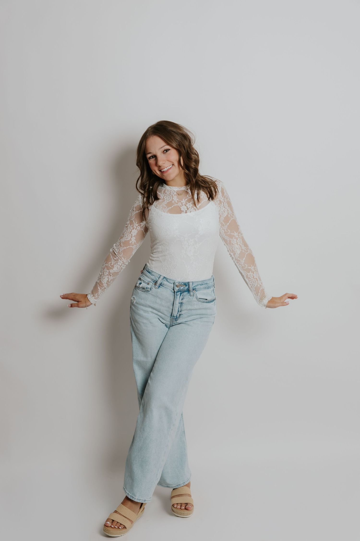 Cheerful woman in white lace top and light wash jeans posing playfully against a white background.