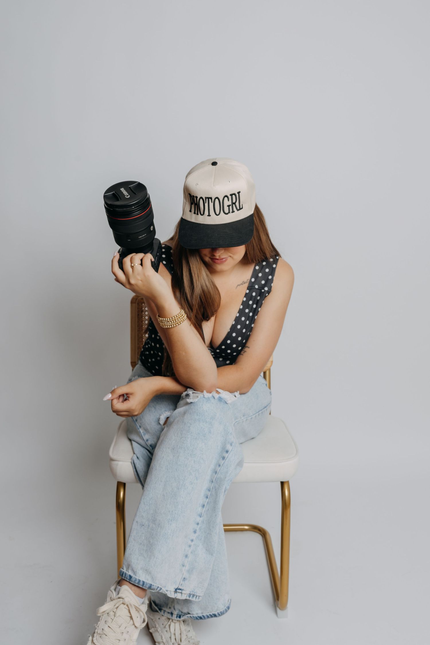 A photographer wearing a PHOTOGRL cap sits on a chair reviewing images on her DSLR camera.