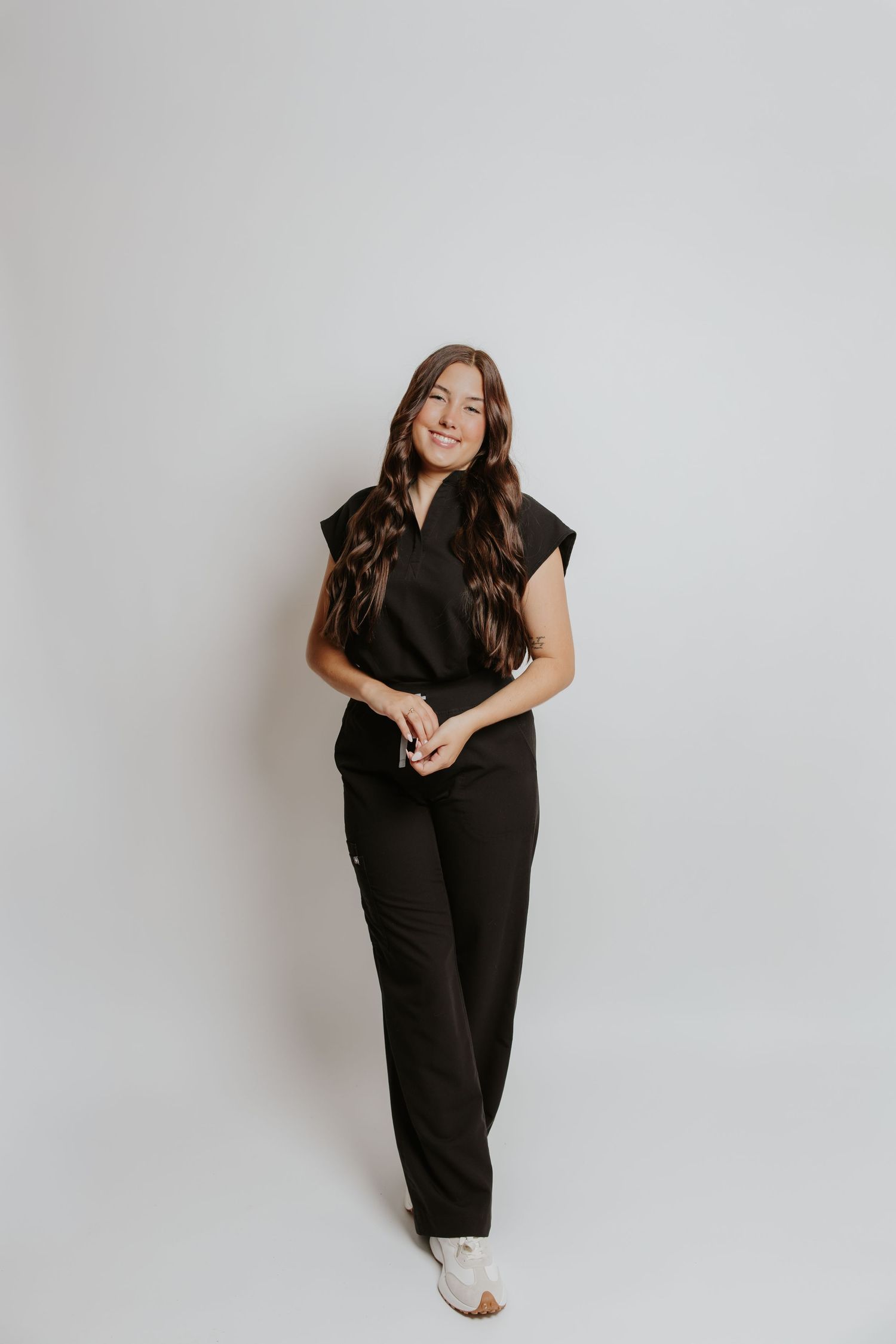 A smiling woman with long brown hair wearing a black jumpsuit and white shoes poses against a light gray background.