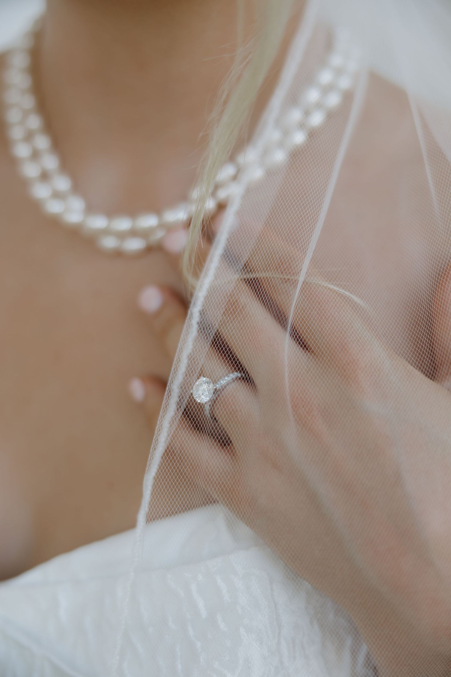 Bride wearing a diamond solitaire engagement ring, pearl necklace, and white wedding dress with veil.