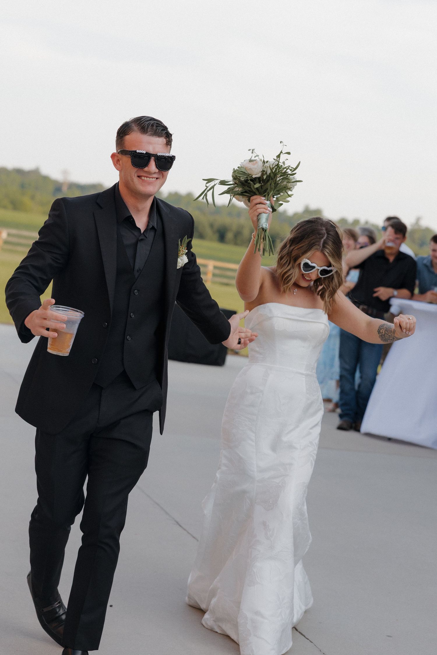 Smiling bride in a white strapless gown and groom in a black suit, both wearing sunglasses, walk joyfully at an outdoor wedding reception.