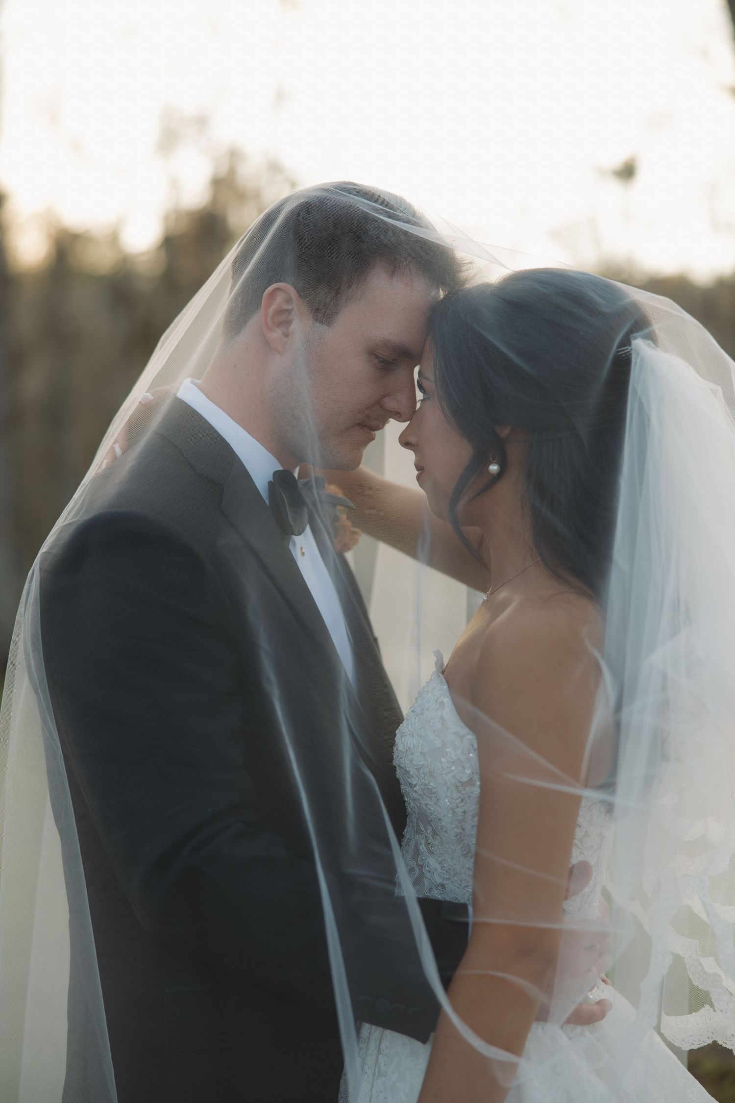 Bride and groom touch foreheads in a tender wedding portrait, wrapped together under a sheer veil in soft golden outdoor light.