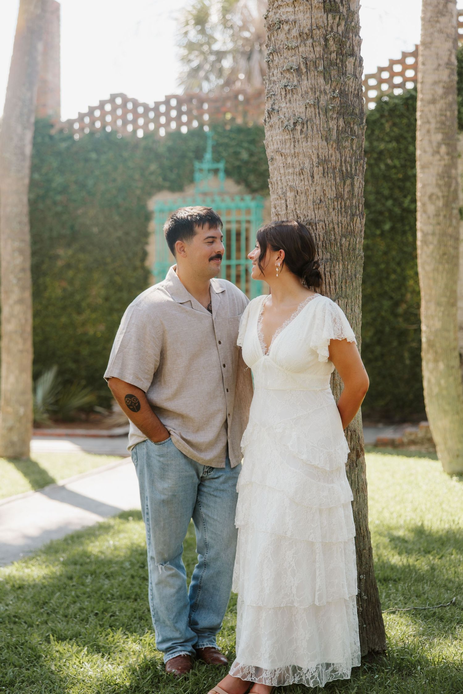 Couple in wedding attire smiling at each other by a palm tree in a lush garden setting.
