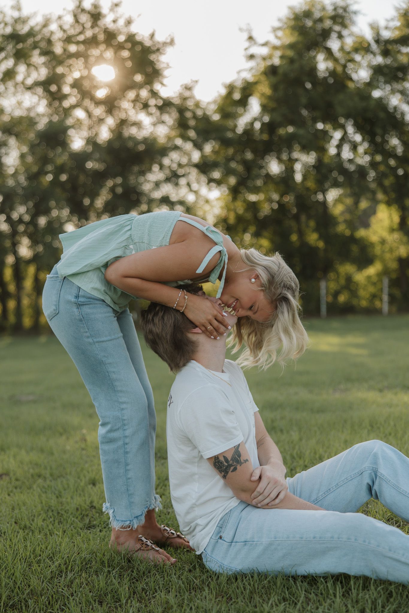 Two women sharing a tender moment on green grass at sunset, one sitting with a tattoo visible, the other leaning in affectionately.
