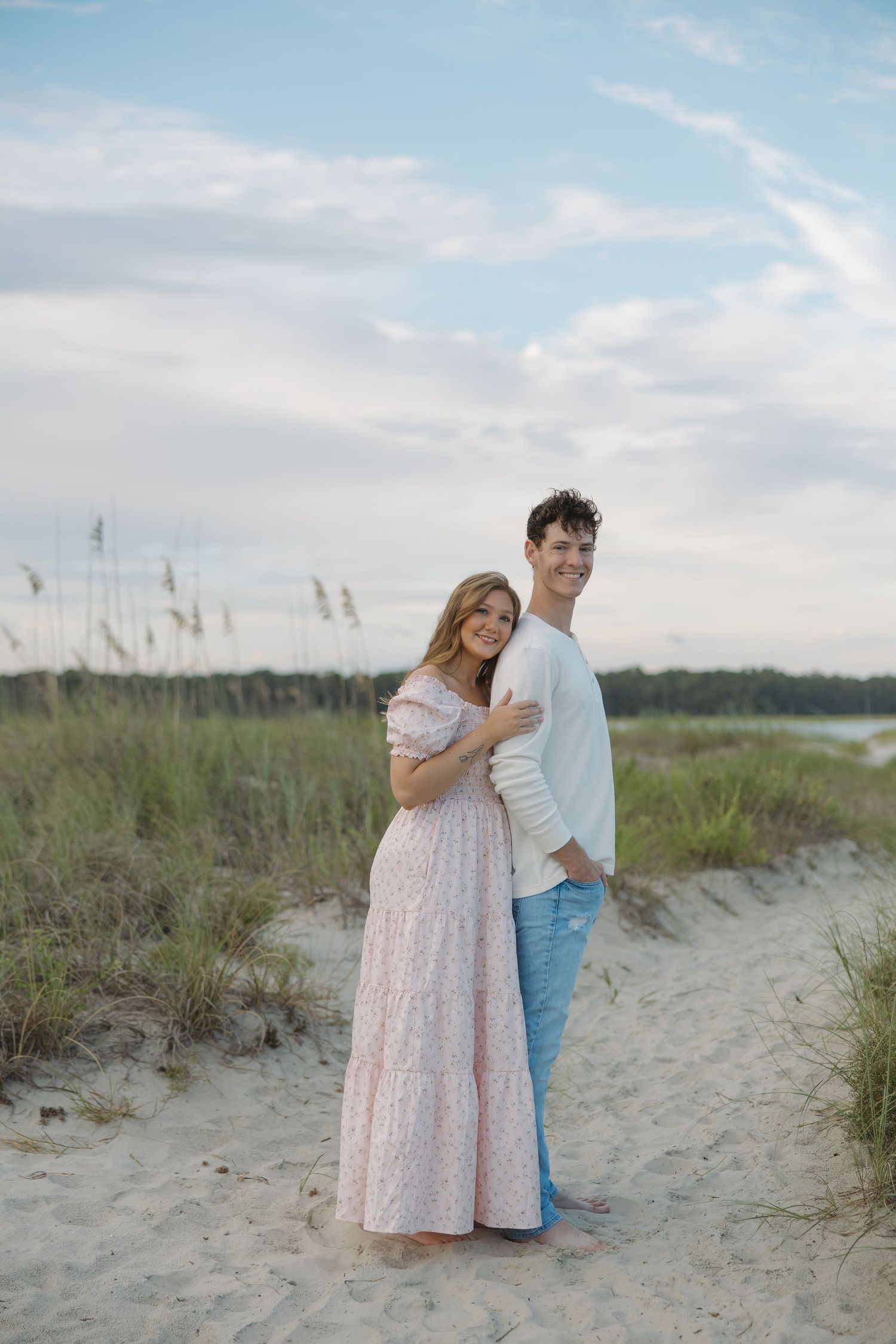 A couple embraces on a sandy beach path, woman in pink floral maxi dress, man in white sweater and jeans, sea oats behind them.