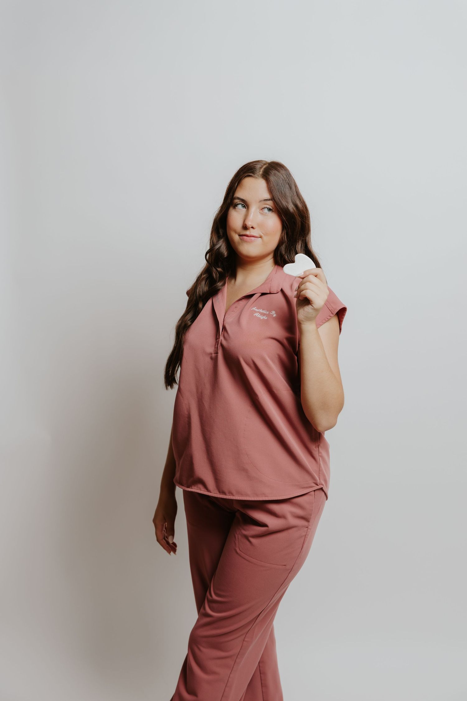 Woman in mauve pink scrubs set poses confidently against a white background, holding a stethoscope.