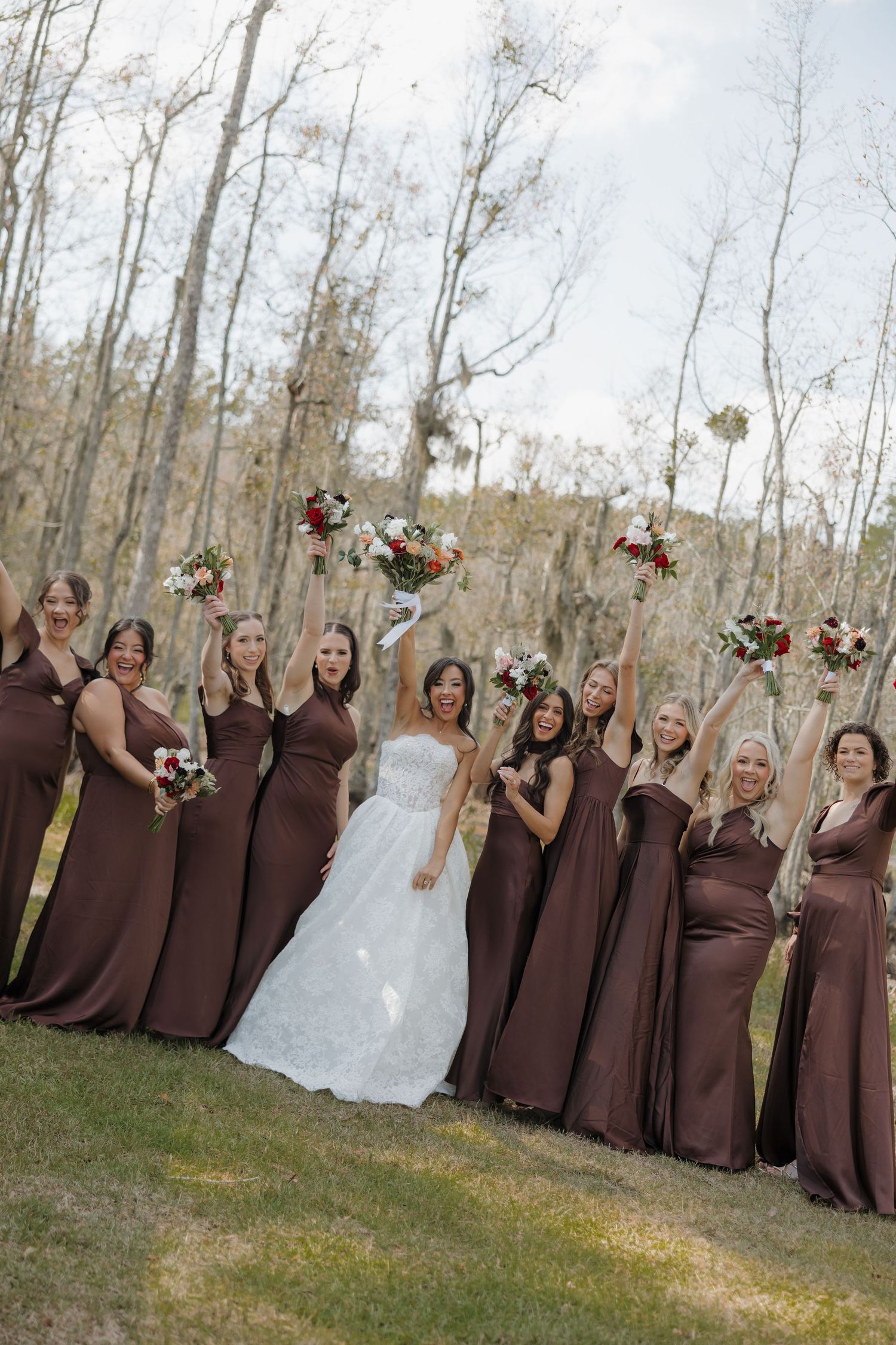 Bride in a white lace gown surrounded by bridesmaids in brown dresses holding bouquets on a grassy lawn near bare trees.