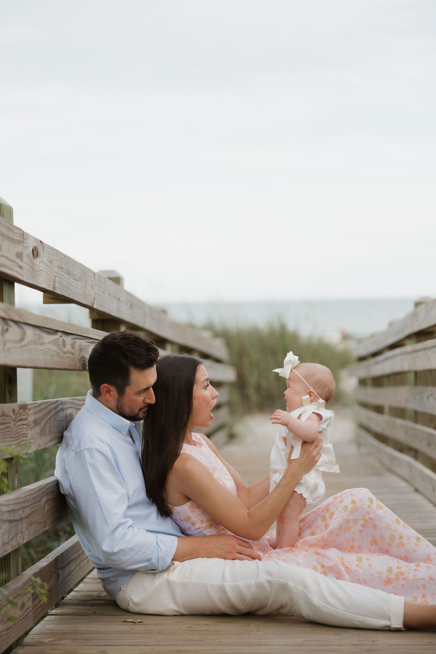 A couple sits on a wooden boardwalk near the beach, lovingly holding their infant daughter.