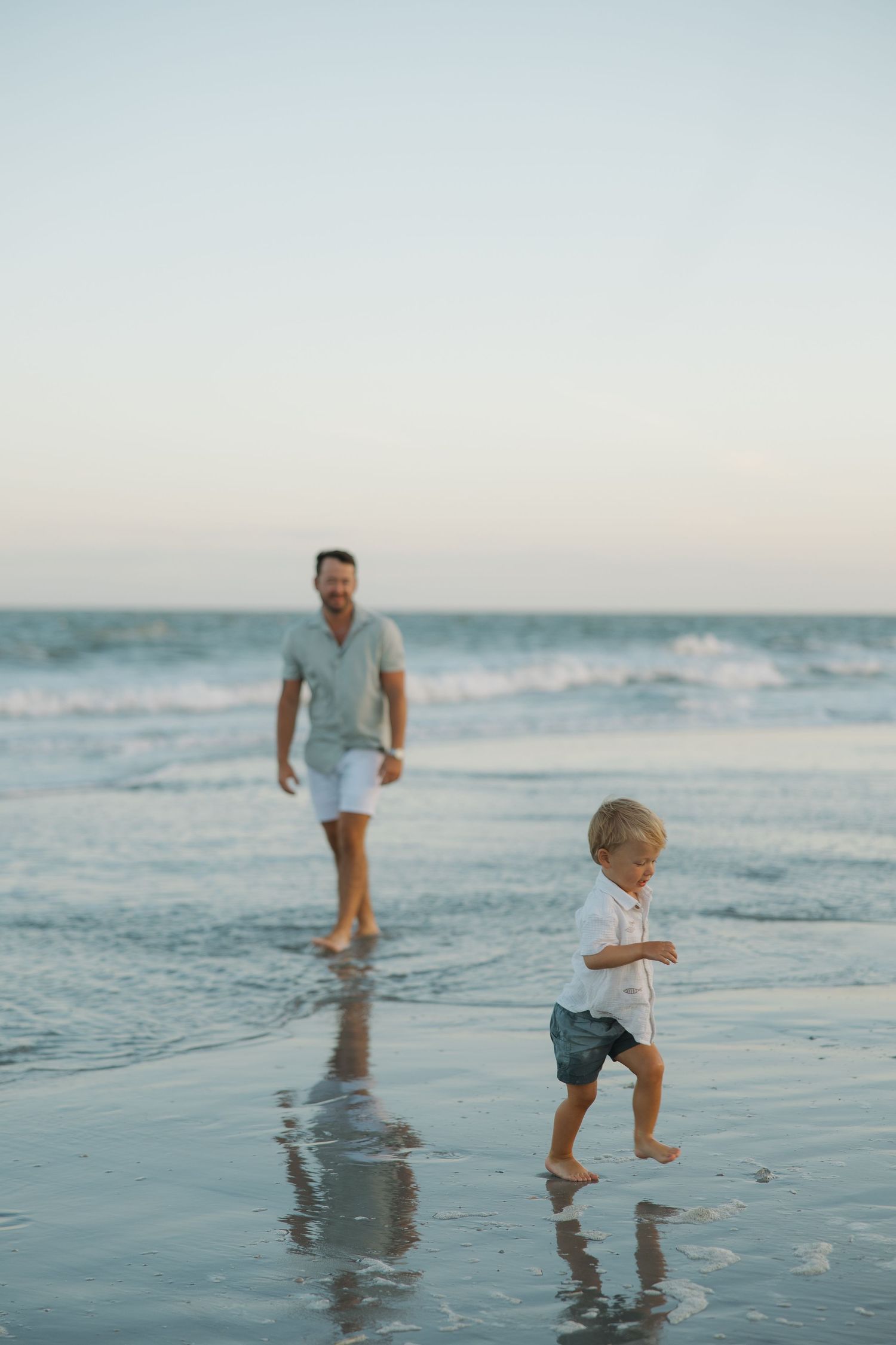 A father and young child playing barefoot on a sandy beach at dusk with ocean waves in the background.