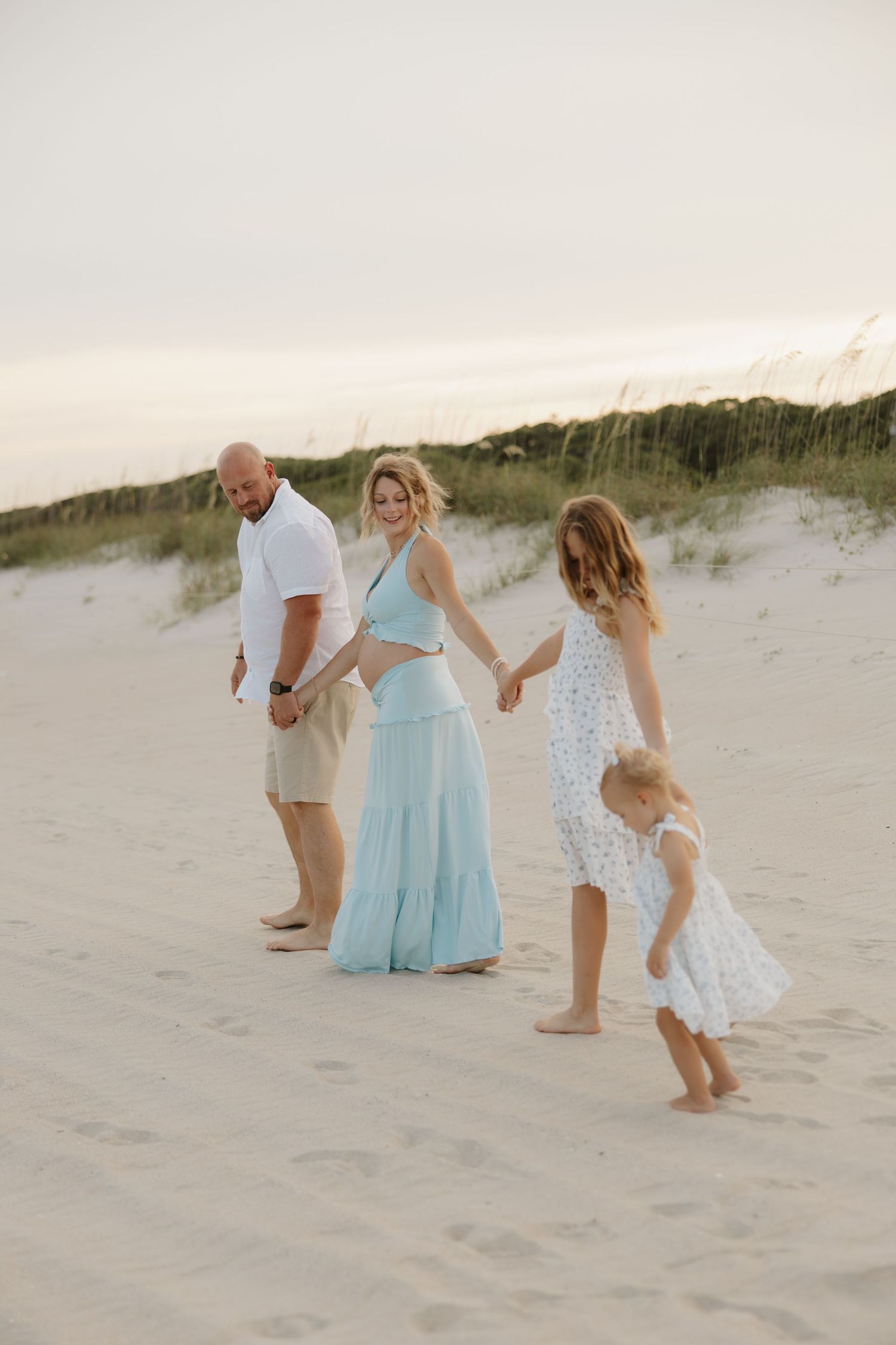 Family of four walking on sandy beach dunes at sunset, wearing light blue and white outfits.