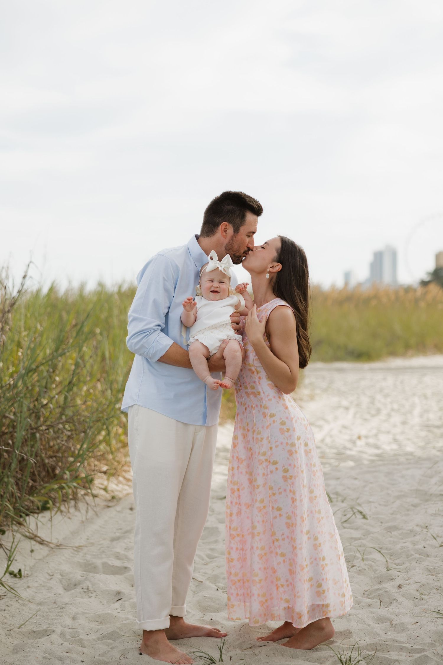 A couple kisses on a sandy beach path while holding their baby dressed in white with a bow headband.