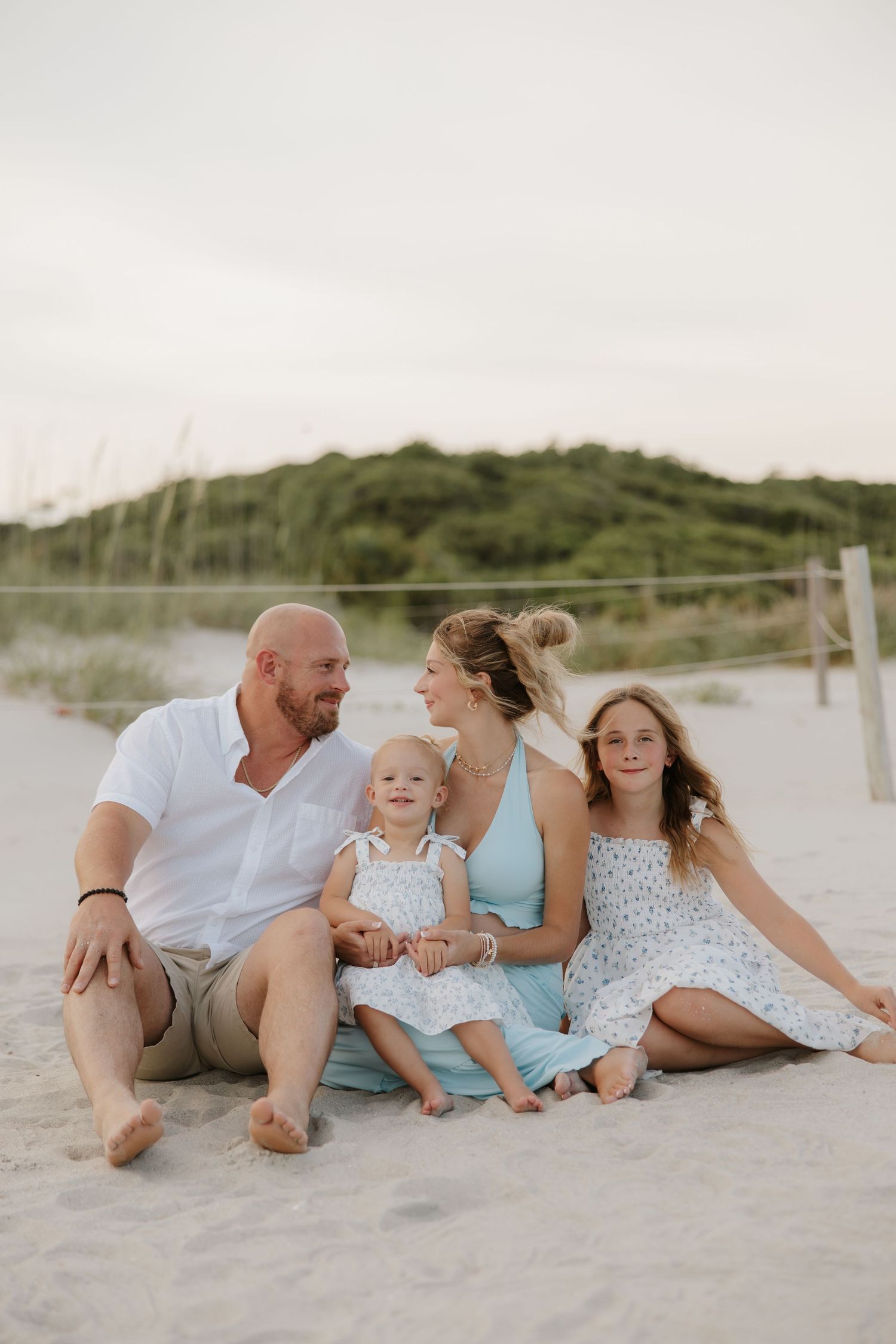 Family of four sitting on sandy beach at sunset, parents and two daughters in light summer outfits, dunes in background.