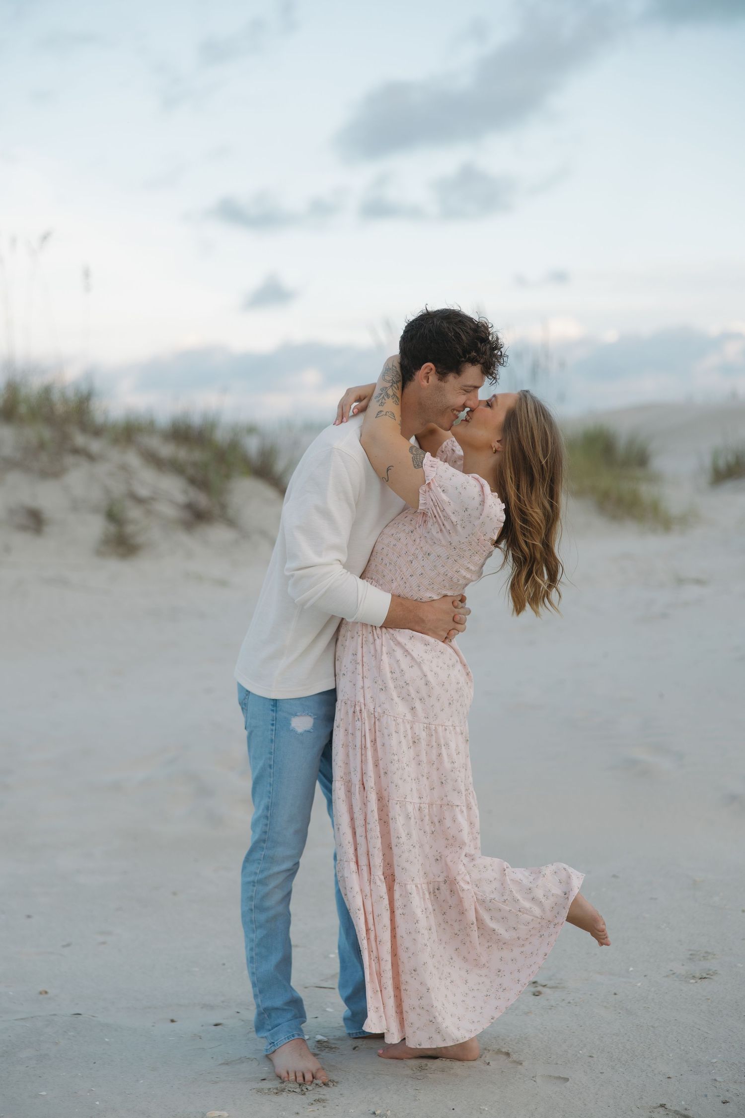 Couple embracing and kissing on a sandy beach at sunset, man in white henley and jeans, woman in flowing pink floral dress.
