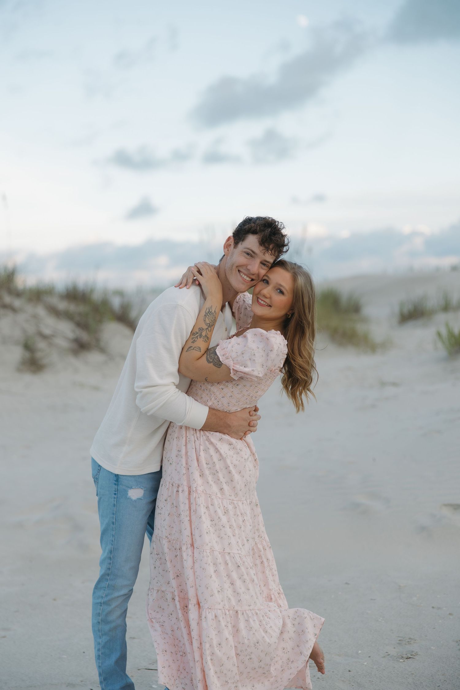 Happy couple embracing on a sandy beach at dusk, woman in pink floral maxi dress and man in white shirt.