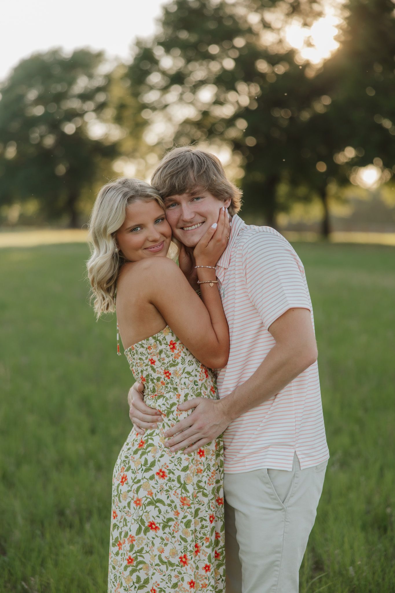 Young couple embracing in a sunlit green field, woman in floral dress smiling over shoulder, man in striped shirt hugging her.