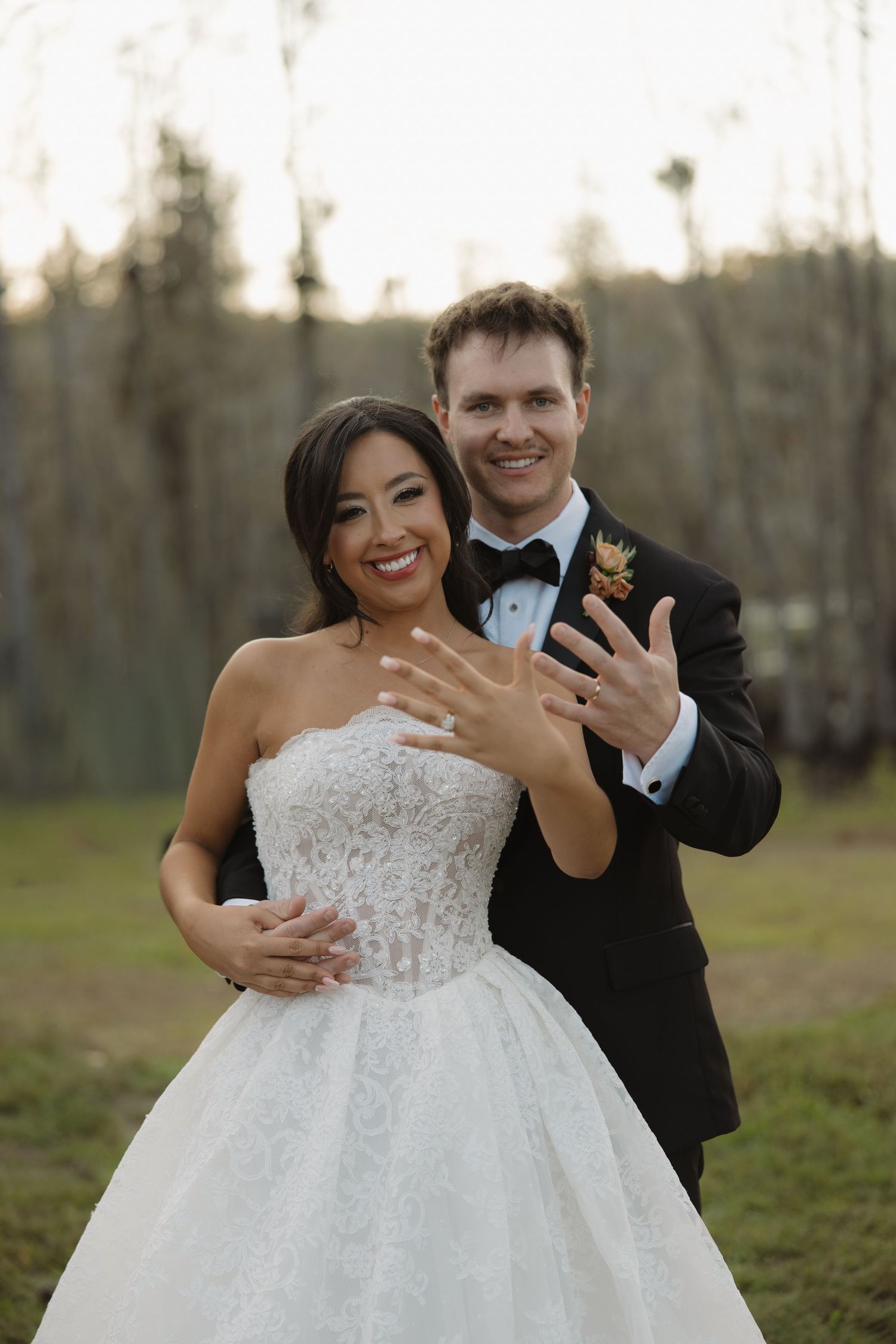 Smiling bride in a strapless lace wedding gown shows her ring while the groom in a black tuxedo and bow tie hugs her from behind.