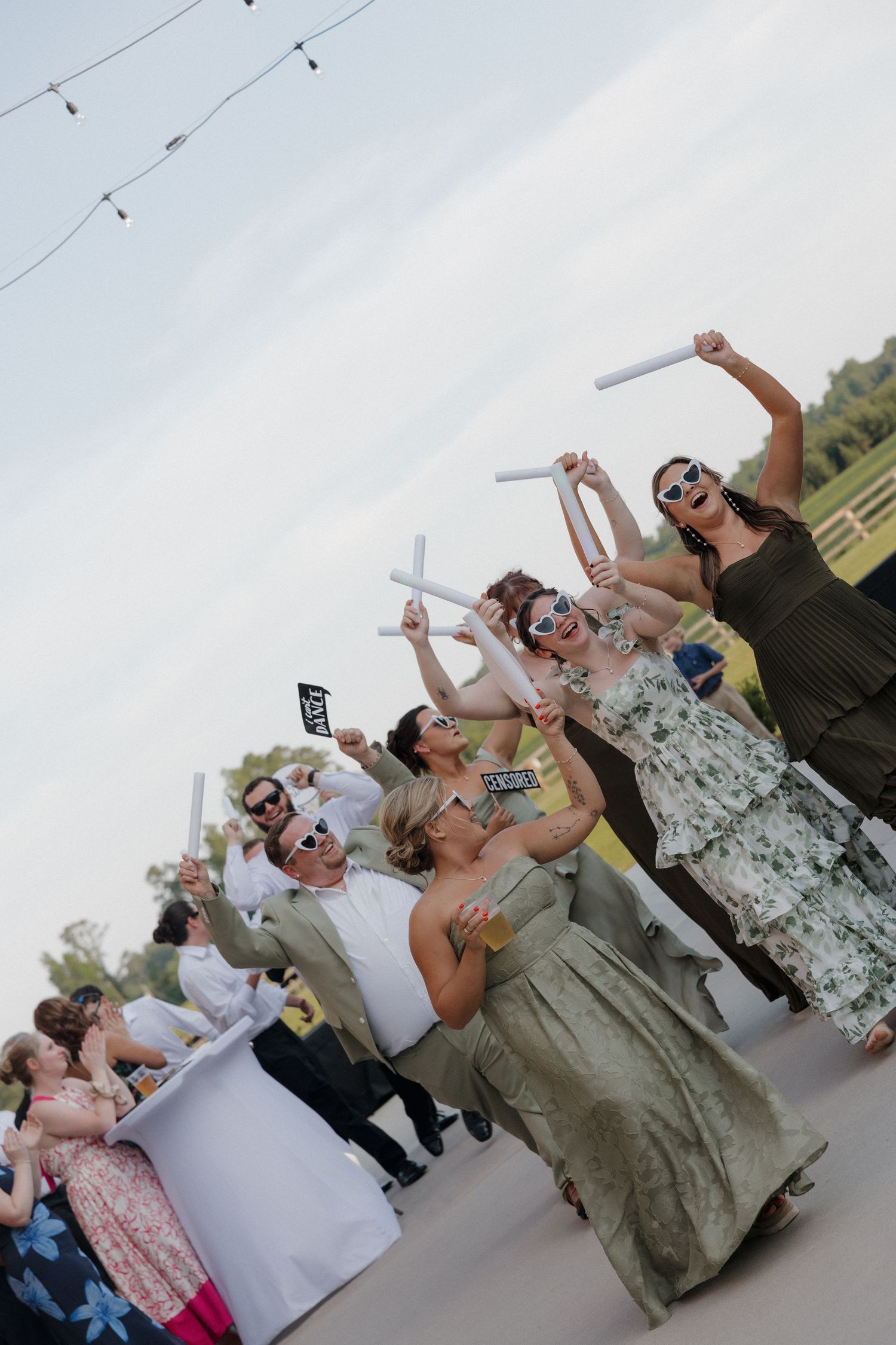 Wedding guests in sunglasses and elegant dresses raise glow sticks and dance outdoors under string lights at a summer reception.