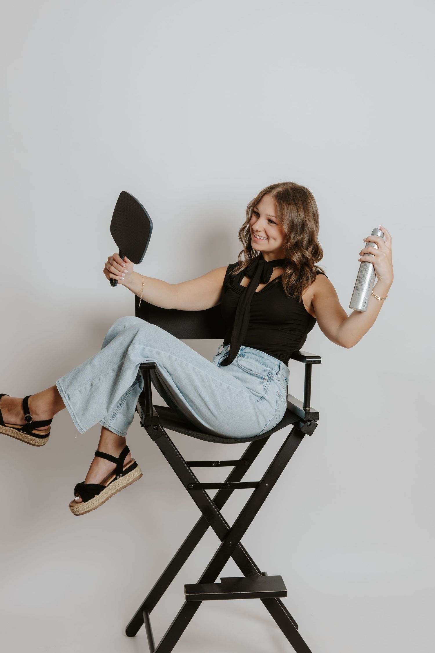 Smiling woman in director chair holds mirror and hairspray can against a white studio background.