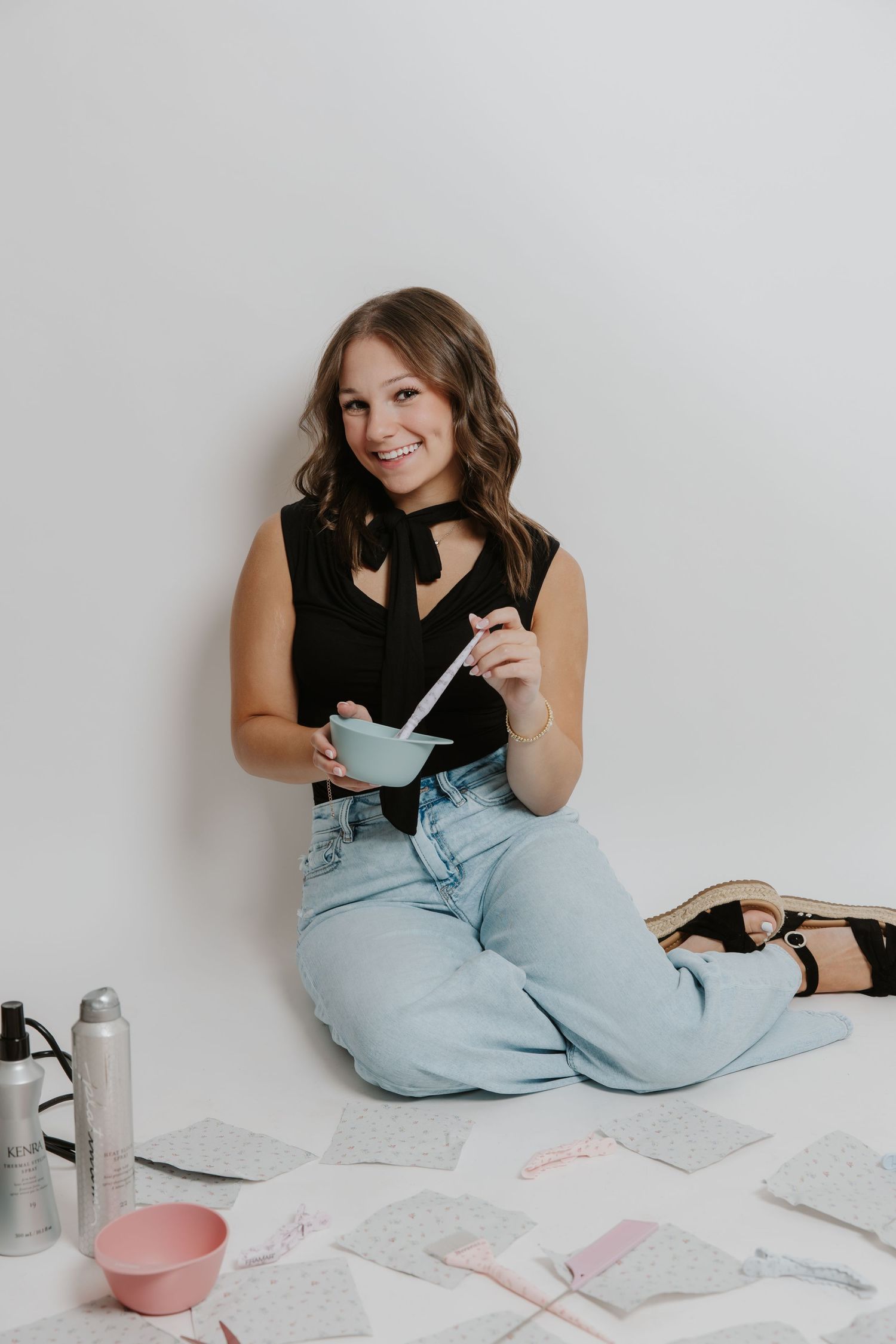Smiling woman sits on floor surrounded by hair tools holding a mixing bowl on white background.