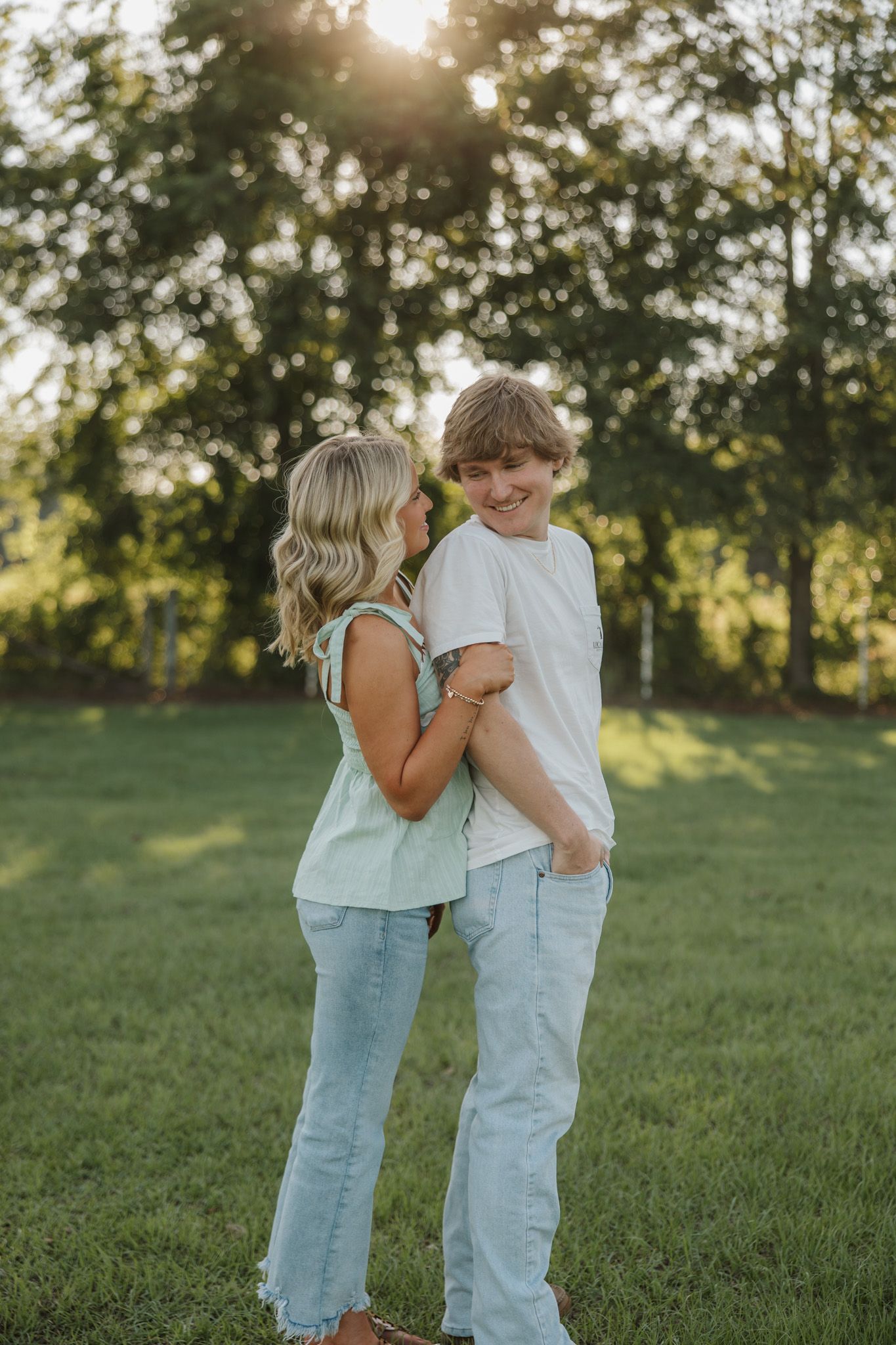 Two teens embrace and smile outdoors on green grass with sunlit trees in the background.