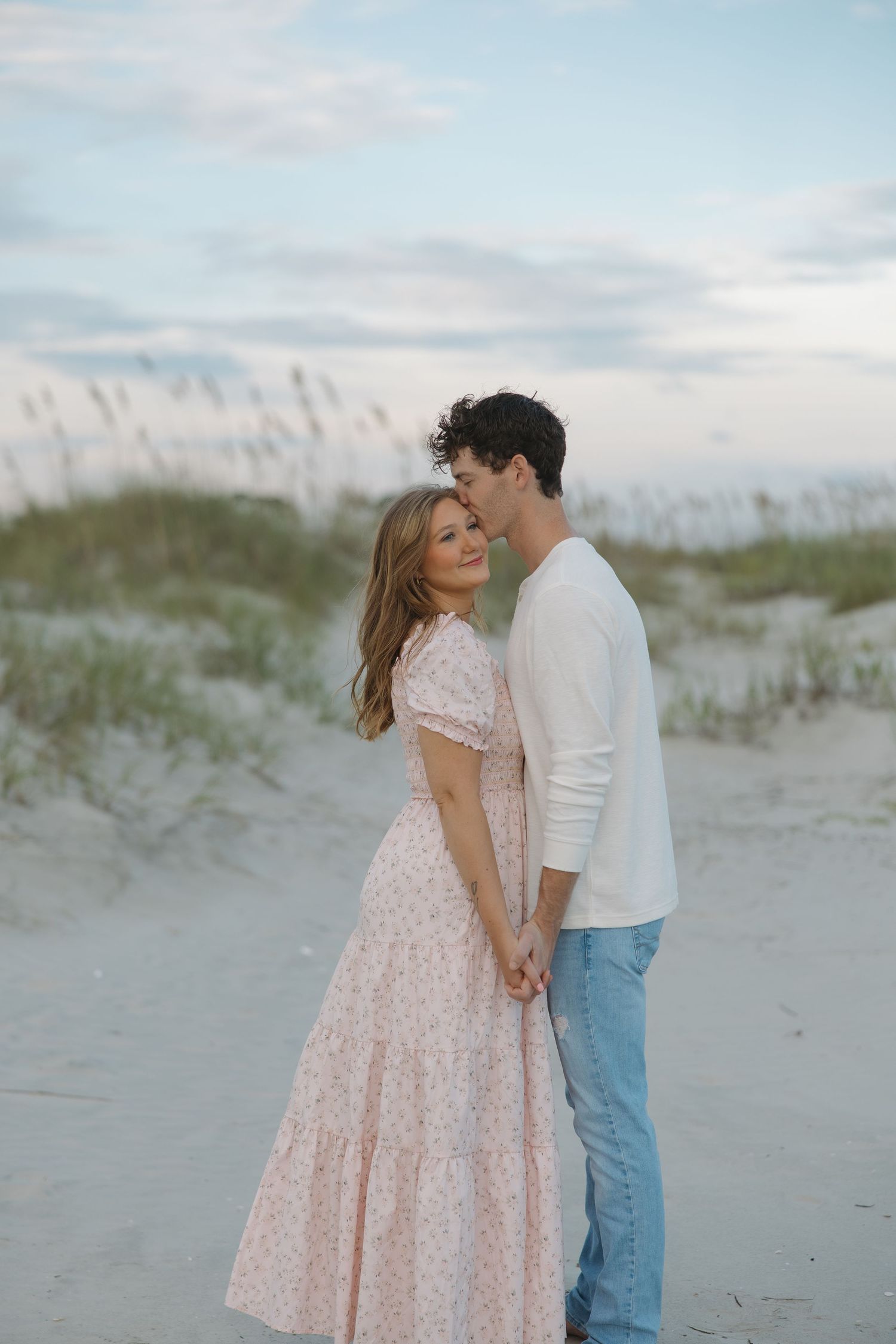 Romantic couple embracing on beach at dusk, woman in pink floral maxi dress, man in white top and jeans.