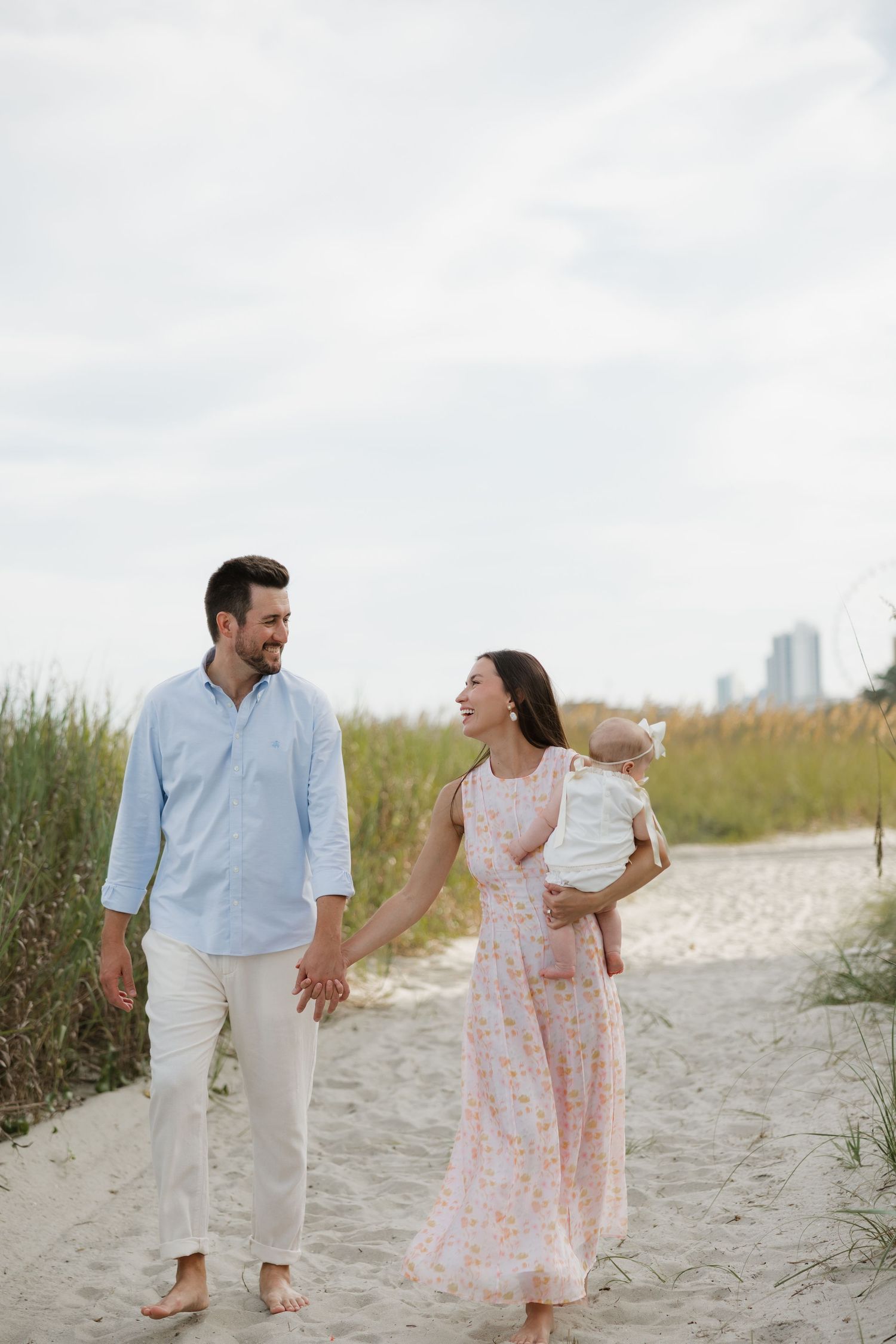 A happy young family walks hand-in-hand on a sandy beach path, with the mother holding their baby.