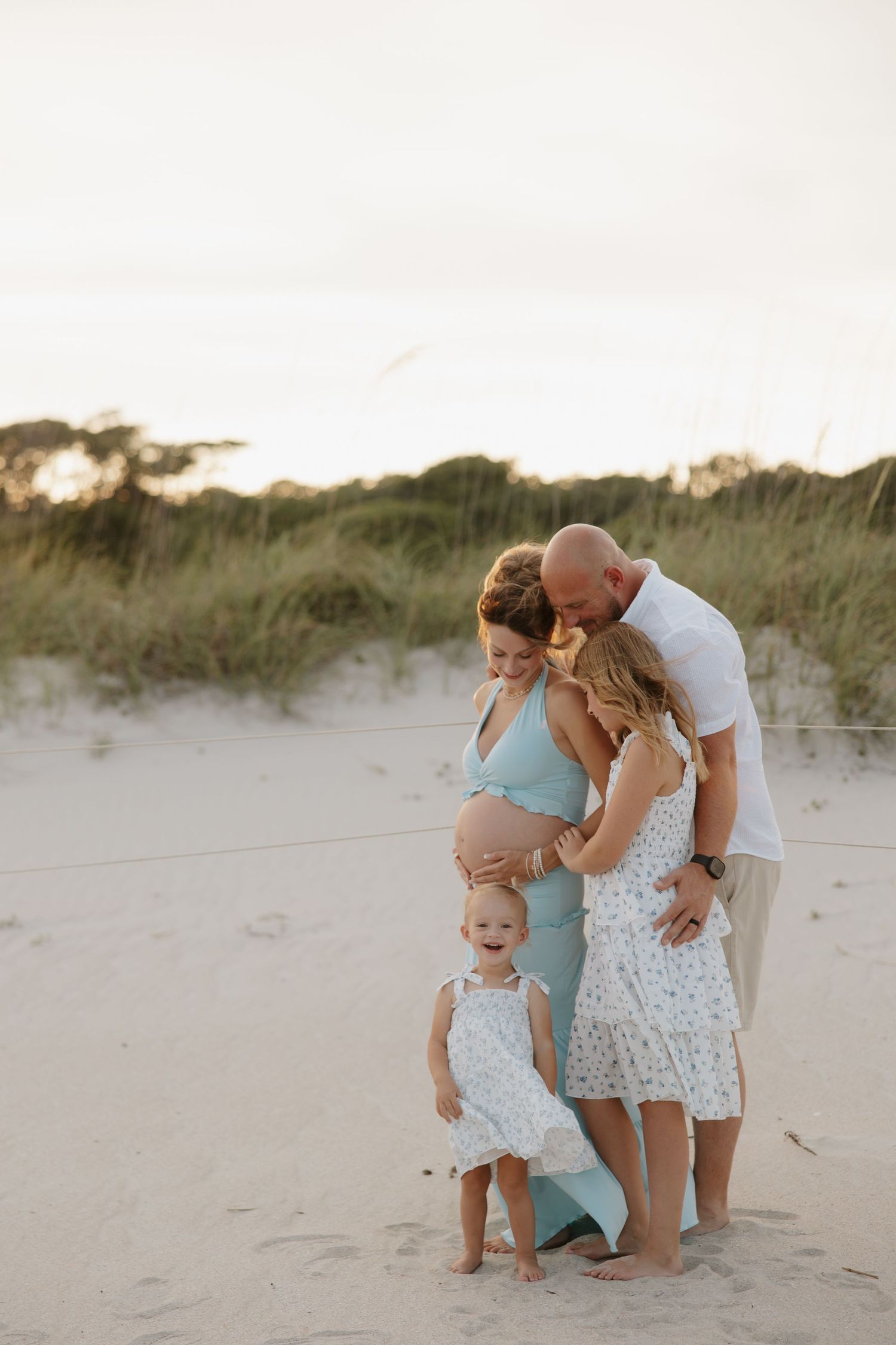 Family maternity beach photo with pregnant mother, father, and two young daughters on sandy dunes at sunset.