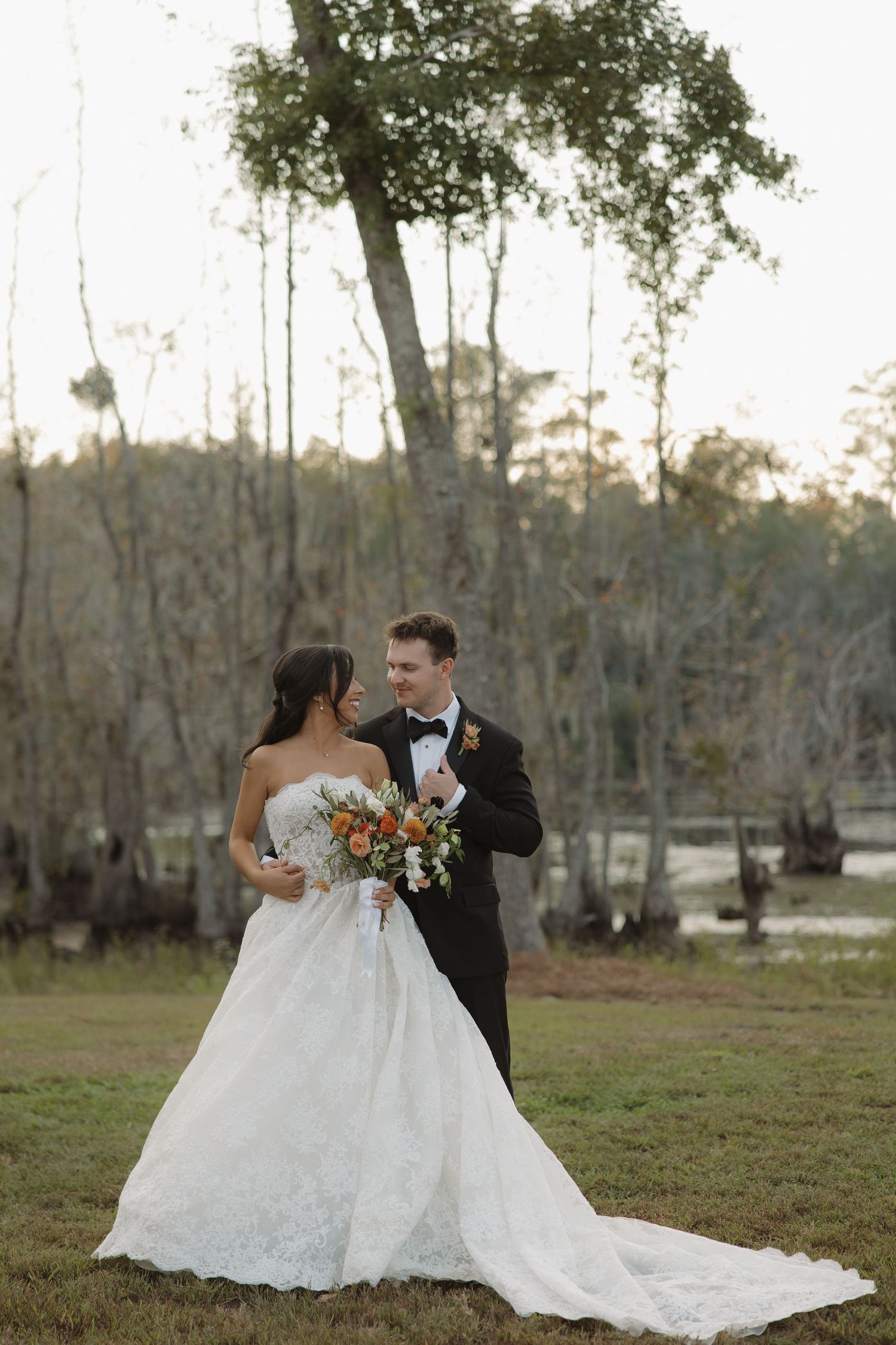Bride in a strapless white lace gown and groom in a black tuxedo hold an orange bouquet on a grassy lawn by a cypress swamp.