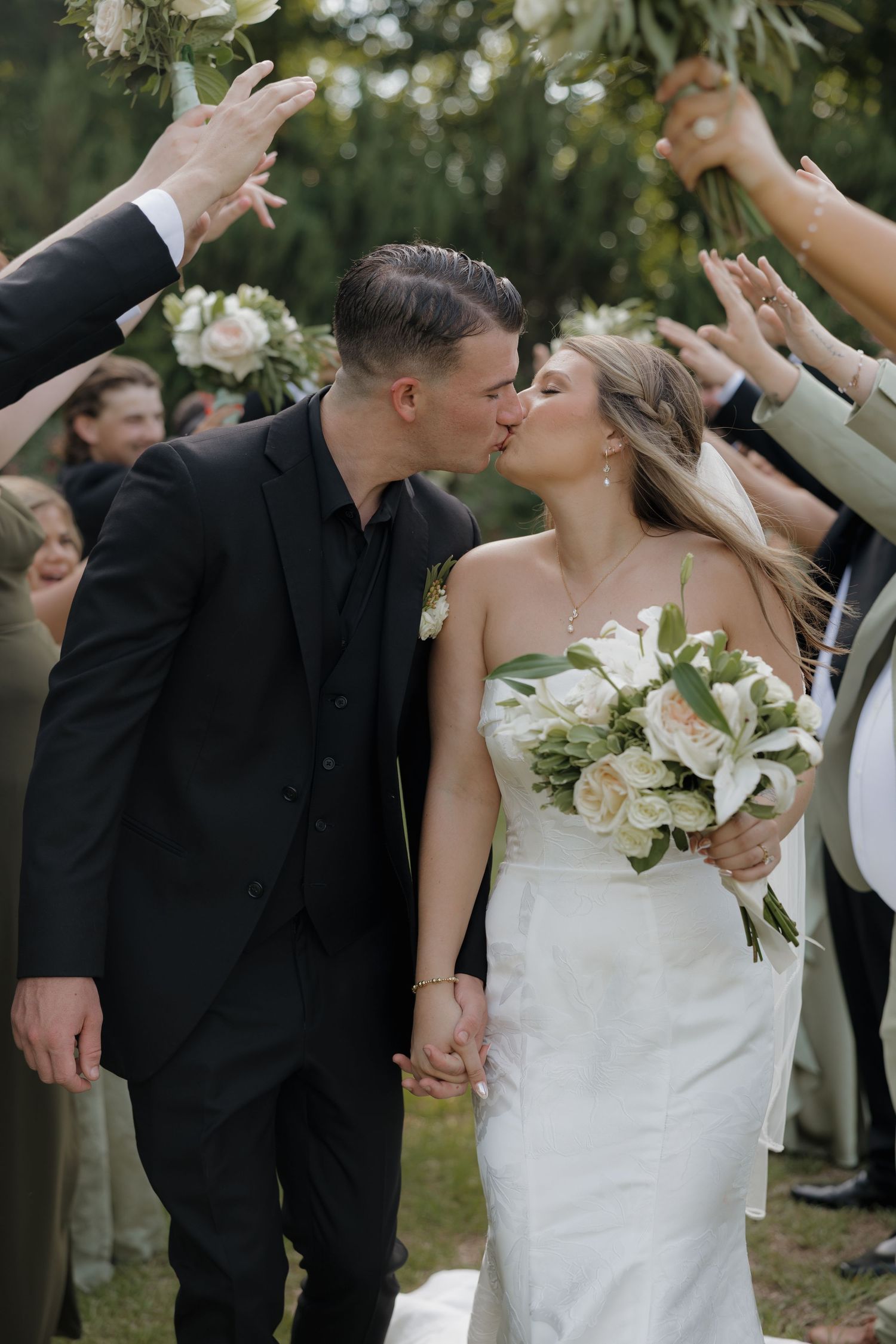 Bride in a white strapless gown holding a white floral bouquet kisses her groom in a black suit as wedding guests cheer with raised hands.