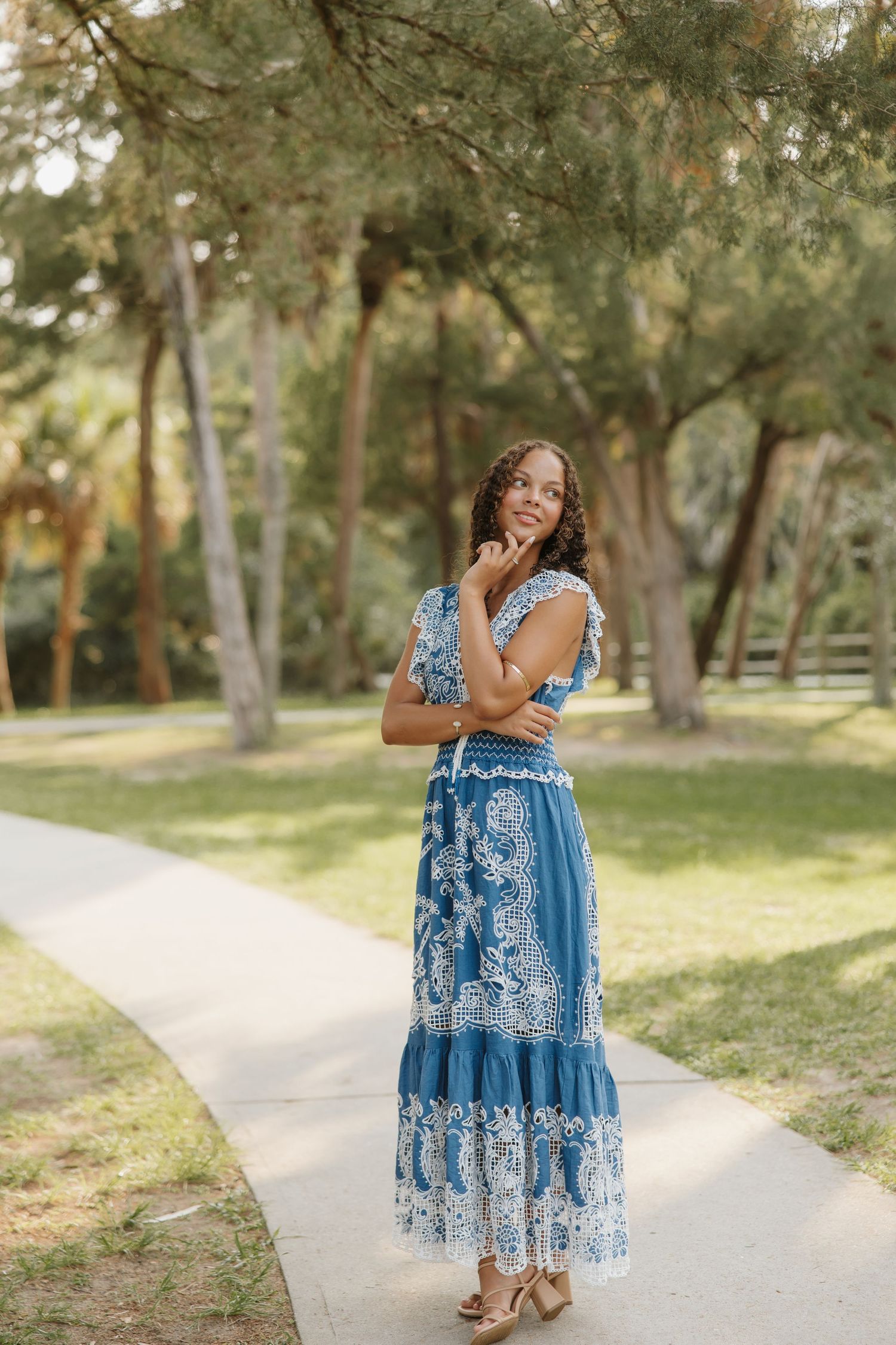Woman in blue and white floral maxi dress standing on a garden path surrounded by lush green trees.