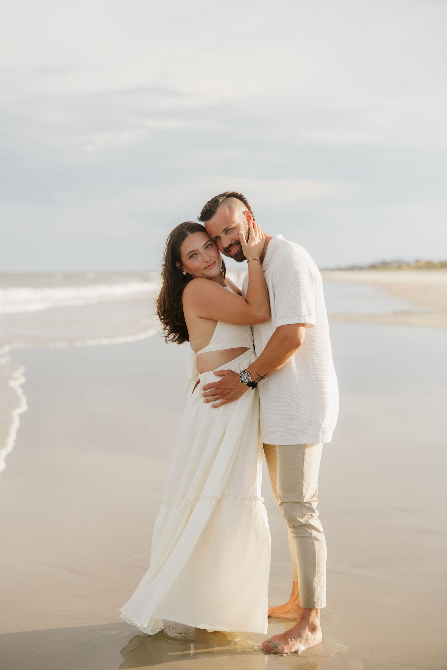Romantic couple hugging at ocean shoreline, woman in white cutout dress and man in white shirt at sunset.
