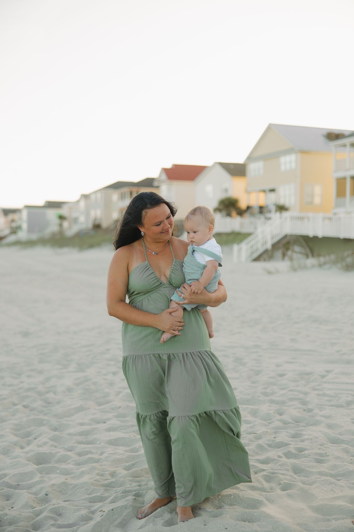 Woman in green maxi dress holding a baby on a sandy beach with coastal houses in the background.