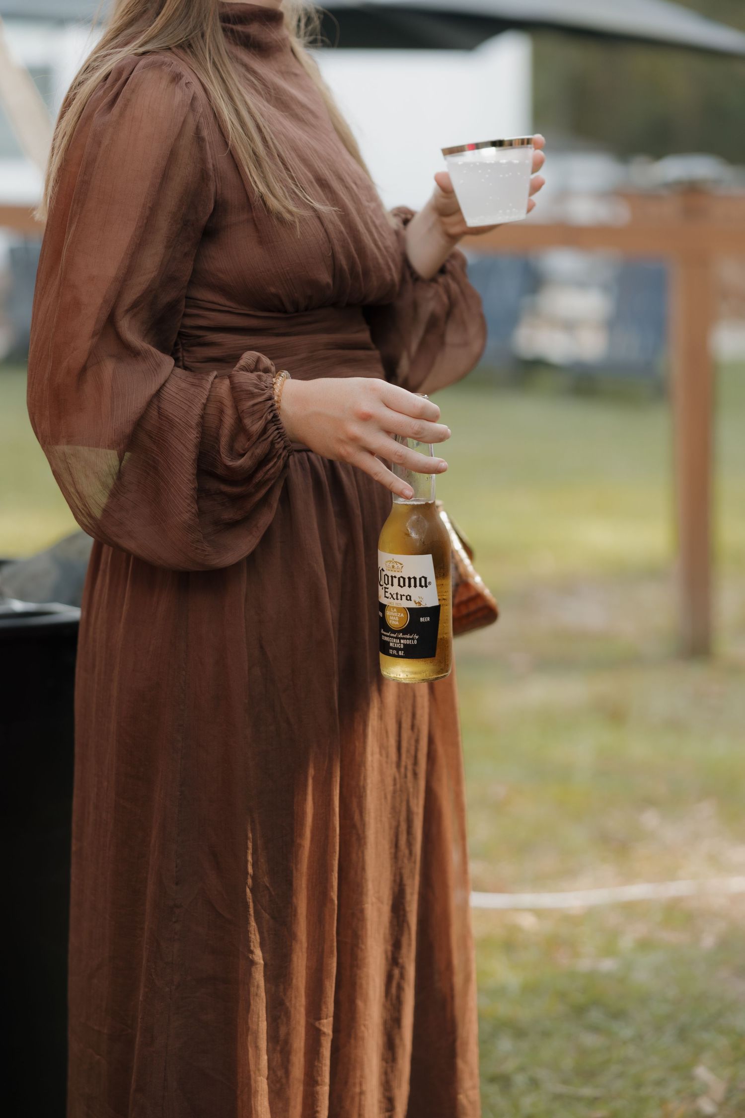 Woman in a flowing brown dress with sheer balloon sleeves holds a Corona Extra beer bottle and a white cup at an outdoor gathering.