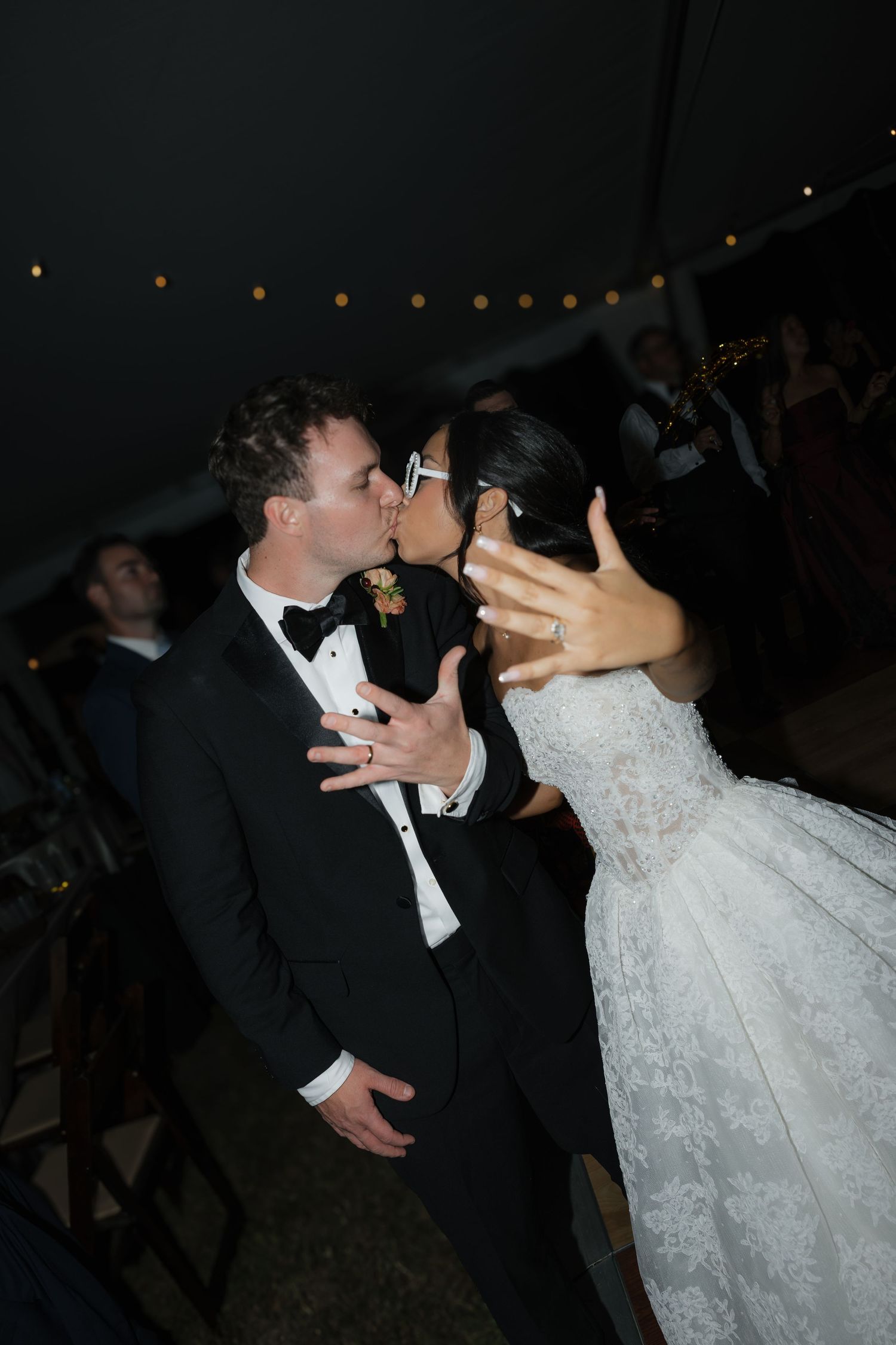 A bride and groom share a kiss at their evening reception as she holds up her hand to show her wedding ring.