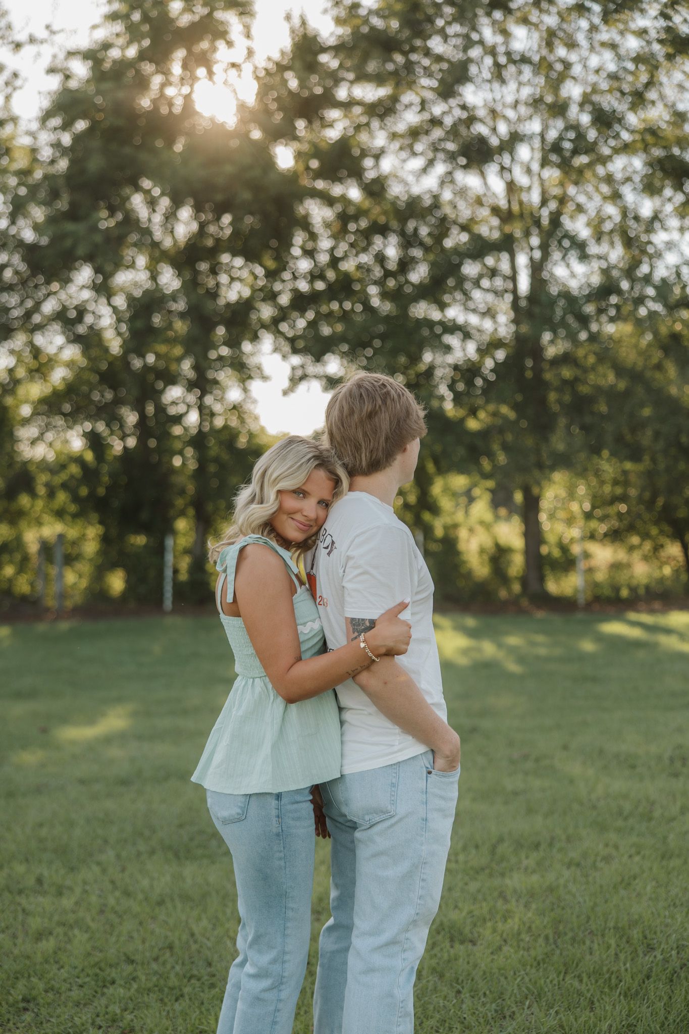 A couple embraces in a sunny green field, woman in mint top and jeans smiling over her shoulder at golden hour.