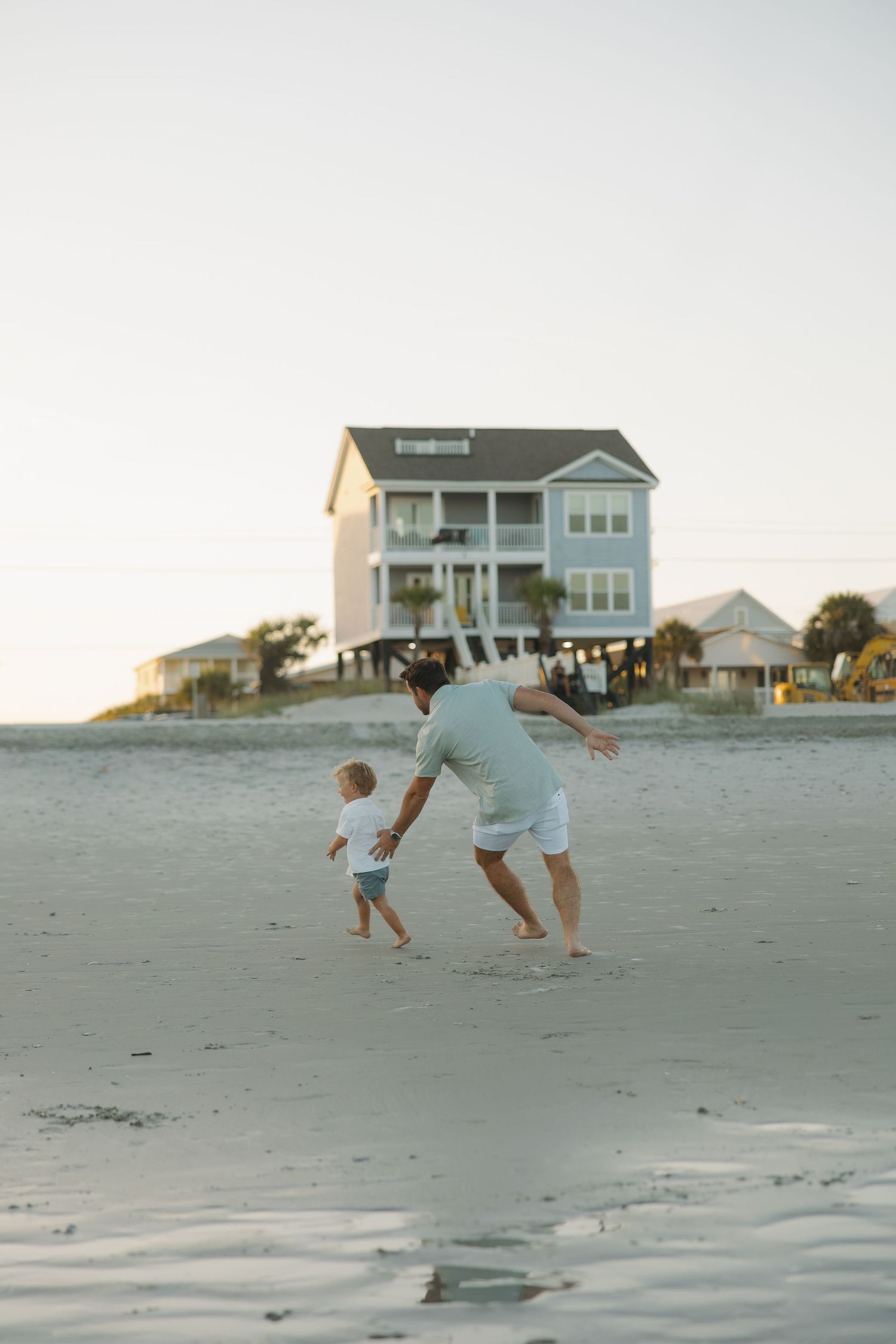 Adult and toddler playing on a sandy beach at sunset with a raised coastal vacation home in the background.