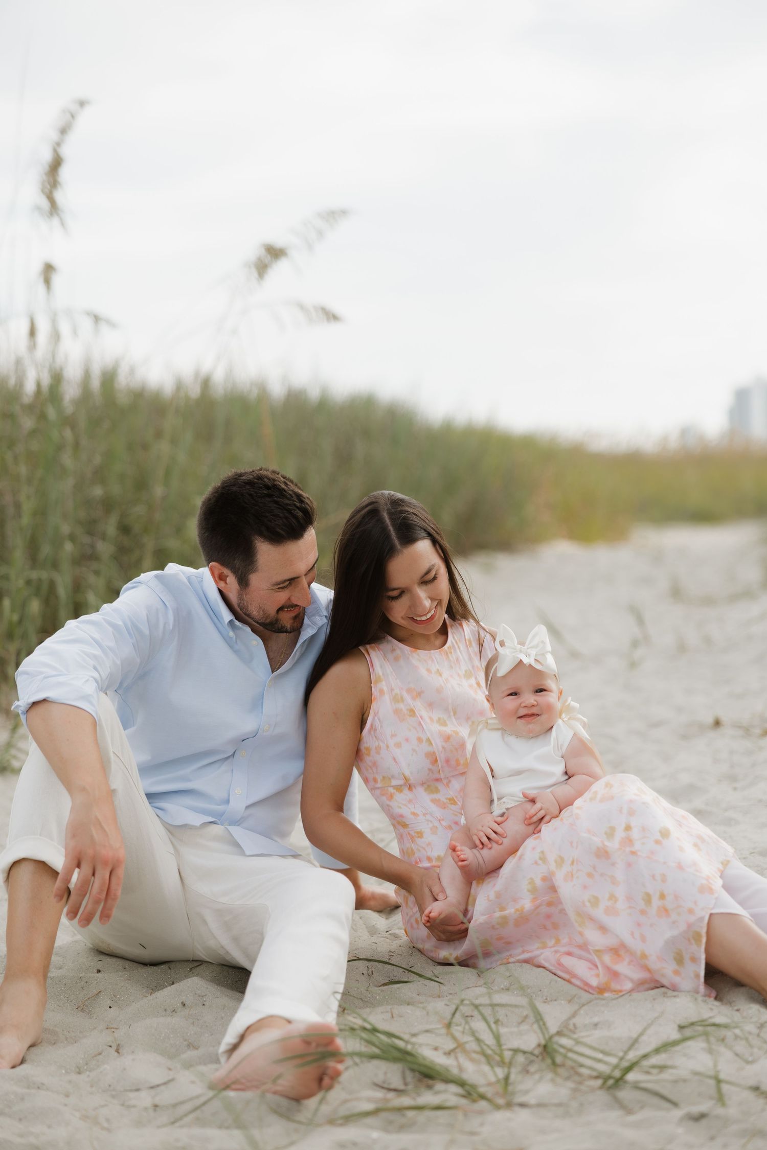 A young family sits on sandy beach dunes, parents smiling at their baby girl in a white outfit and bow headband.