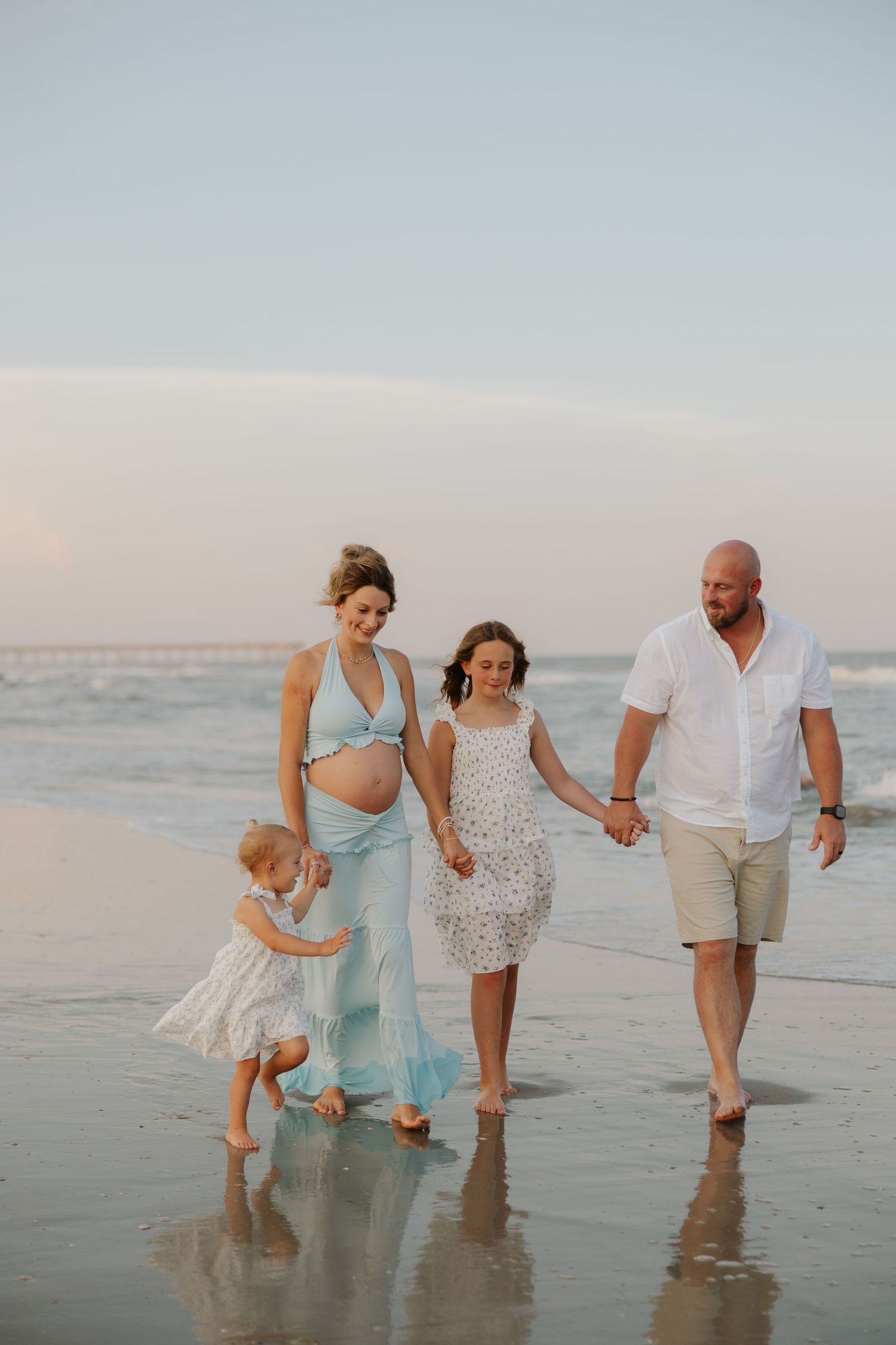 Pregnant woman in blue dress walks on beach at sunset with husband and two young daughters.