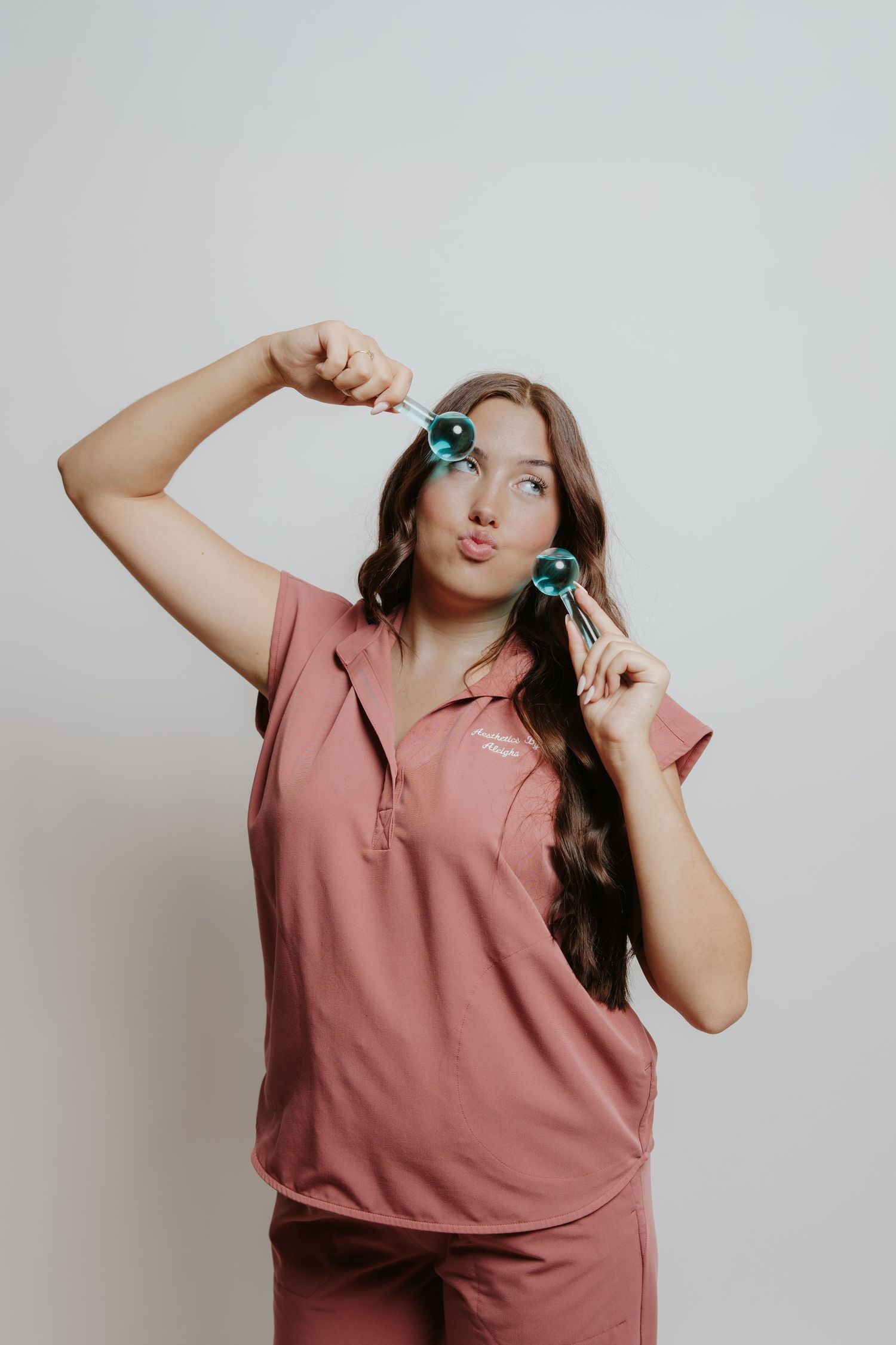 Woman in pink scrubs holding teal facial ice globes, making a kissing face against a light gray background.