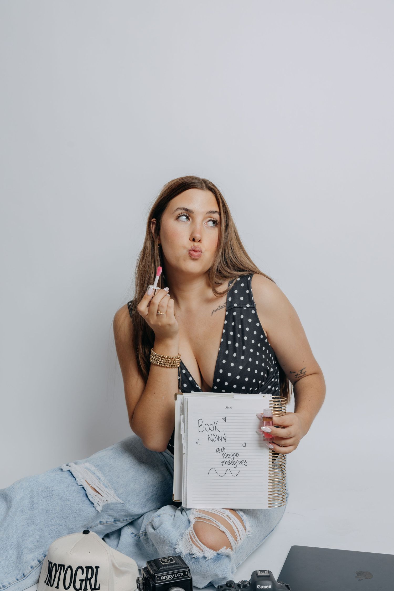 Woman in polka dot top holding lipstick and spiral notebook with book now text on white background.