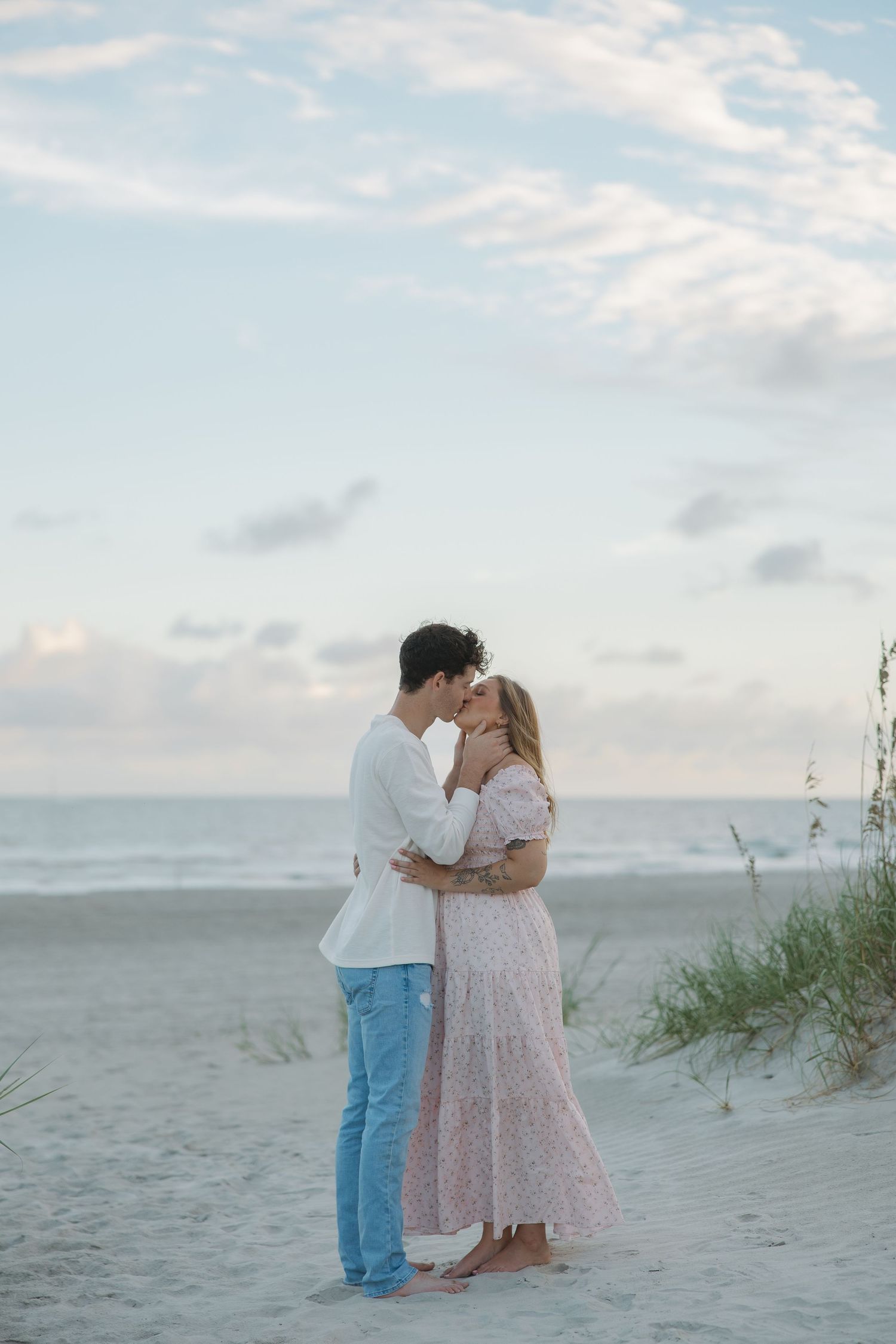 Couple kissing on sandy beach at sunset, woman in floral dress, man in white shirt.
