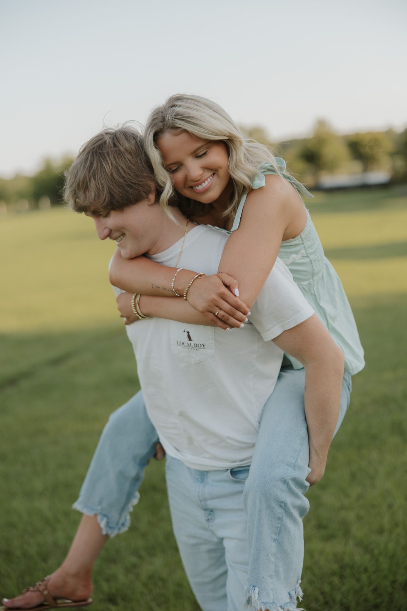 A smiling woman gets a piggyback ride from a man in a white tee on a green grass field.