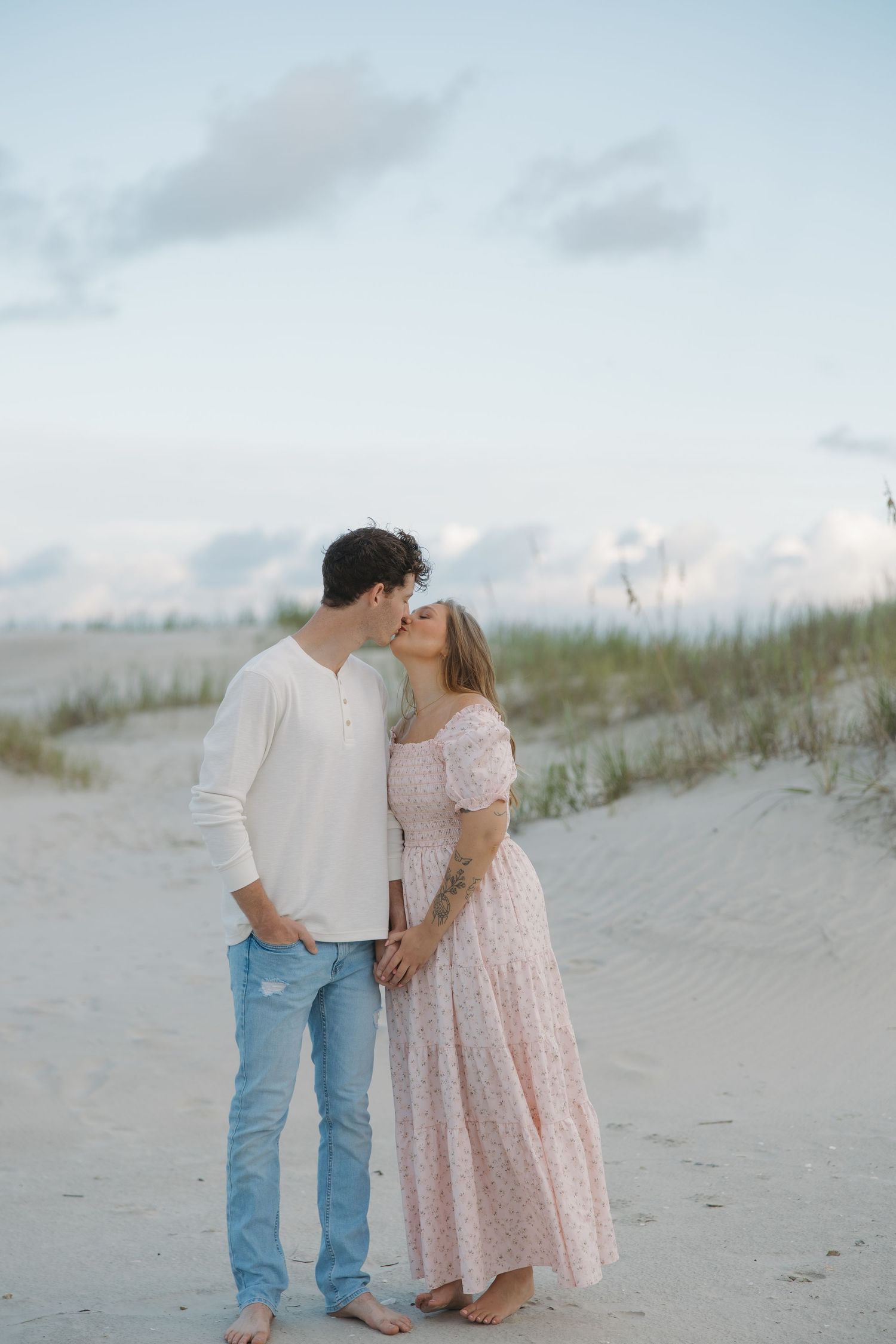 Couple sharing a romantic kiss on a sandy beach at dusk, man in white shirt and jeans, woman in pink floral maxi dress.