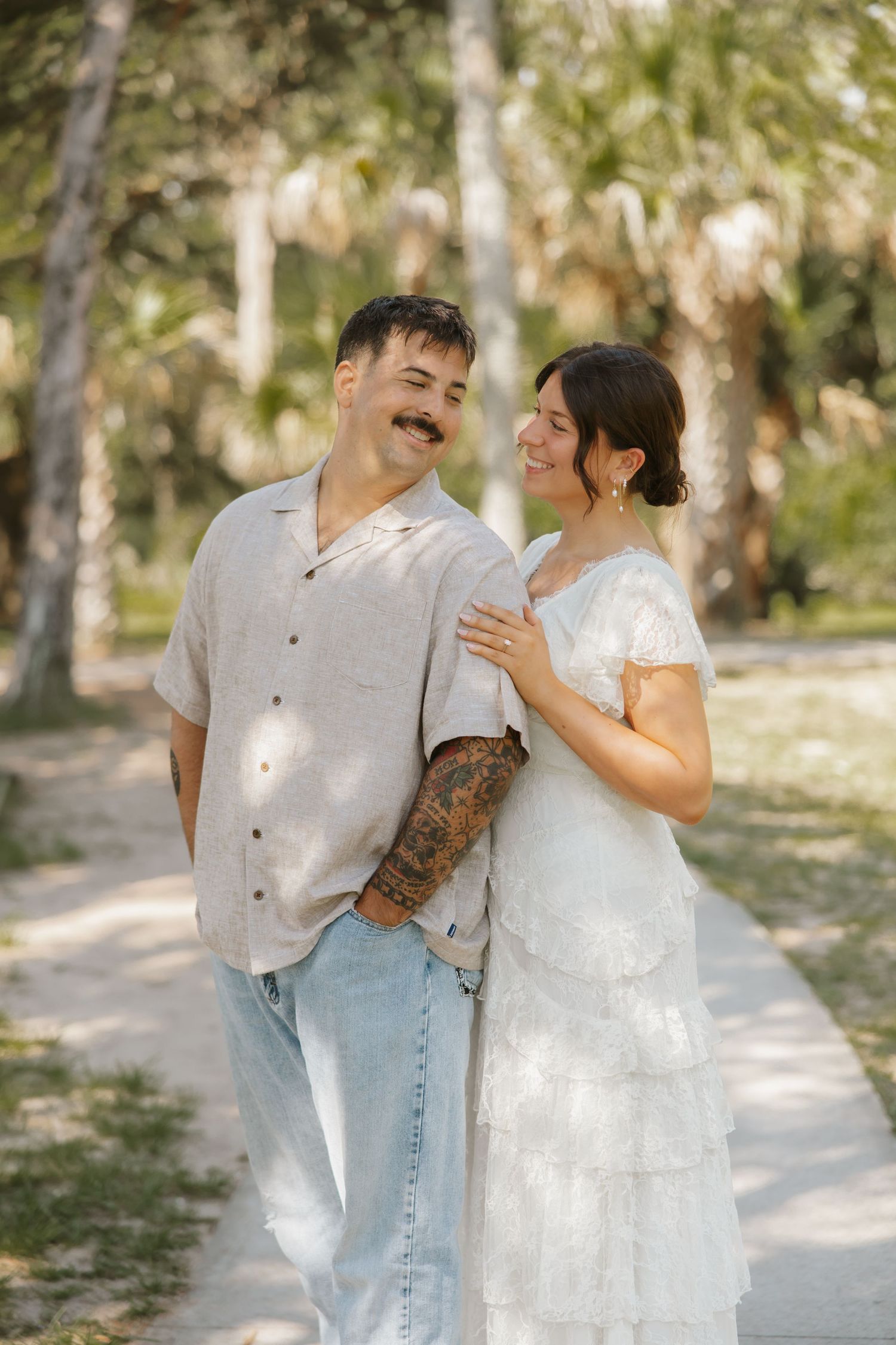 A smiling couple poses on a sunlit path surrounded by palm trees, the woman in a white lace dress and man in a linen shirt with tattoos.