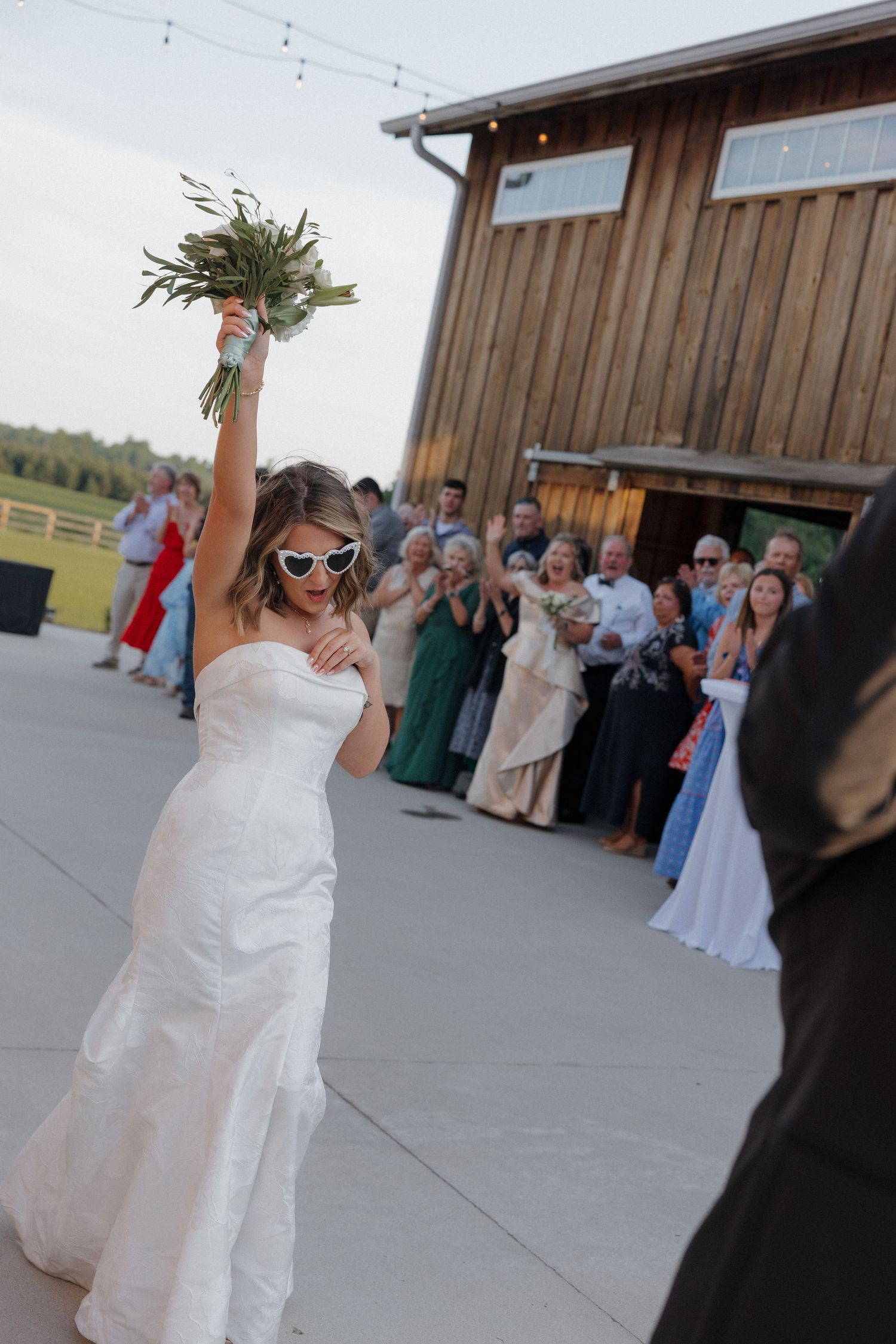 Bride in white sunglasses and a strapless gown raises her bouquet to toss it as guests cheer outside a rustic wooden barn venue.